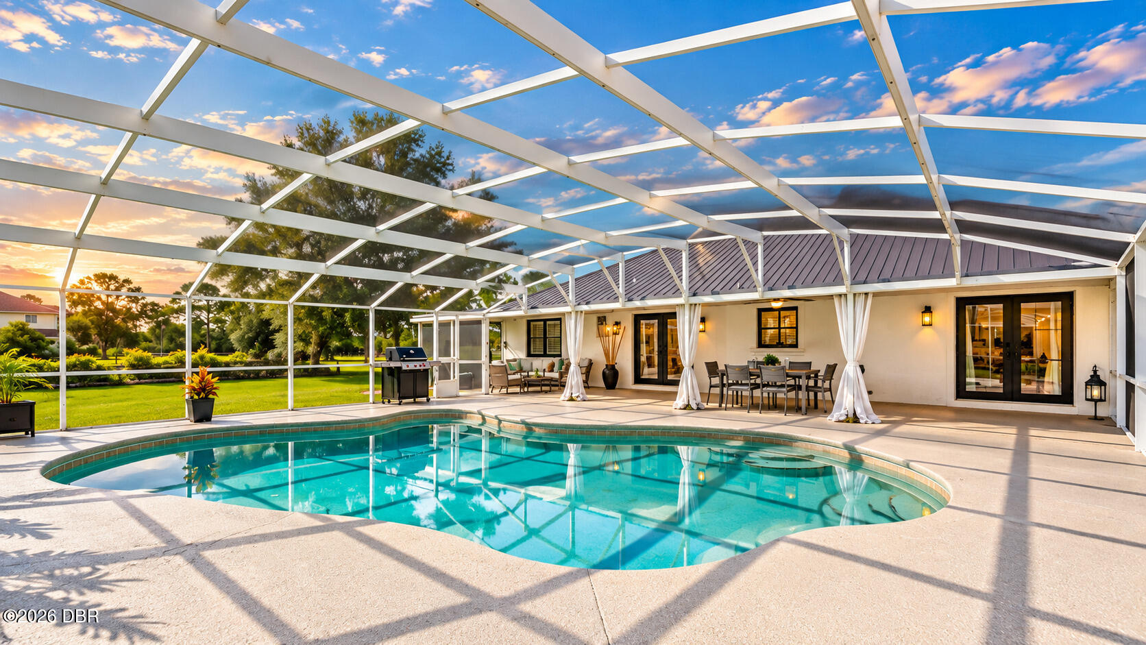 Curved swimming pool beneath a screened enclosure, with a furnished patio and lawn beyond.