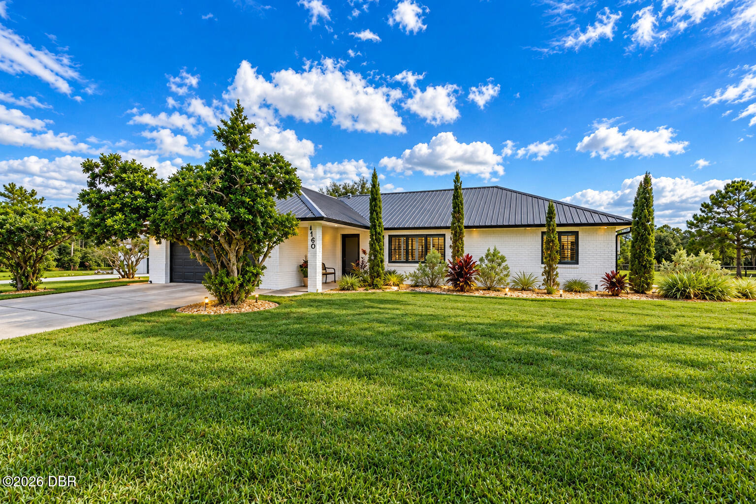Single-story white brick home with a dark metal roof, landscaped beds, and a broad green lawn.