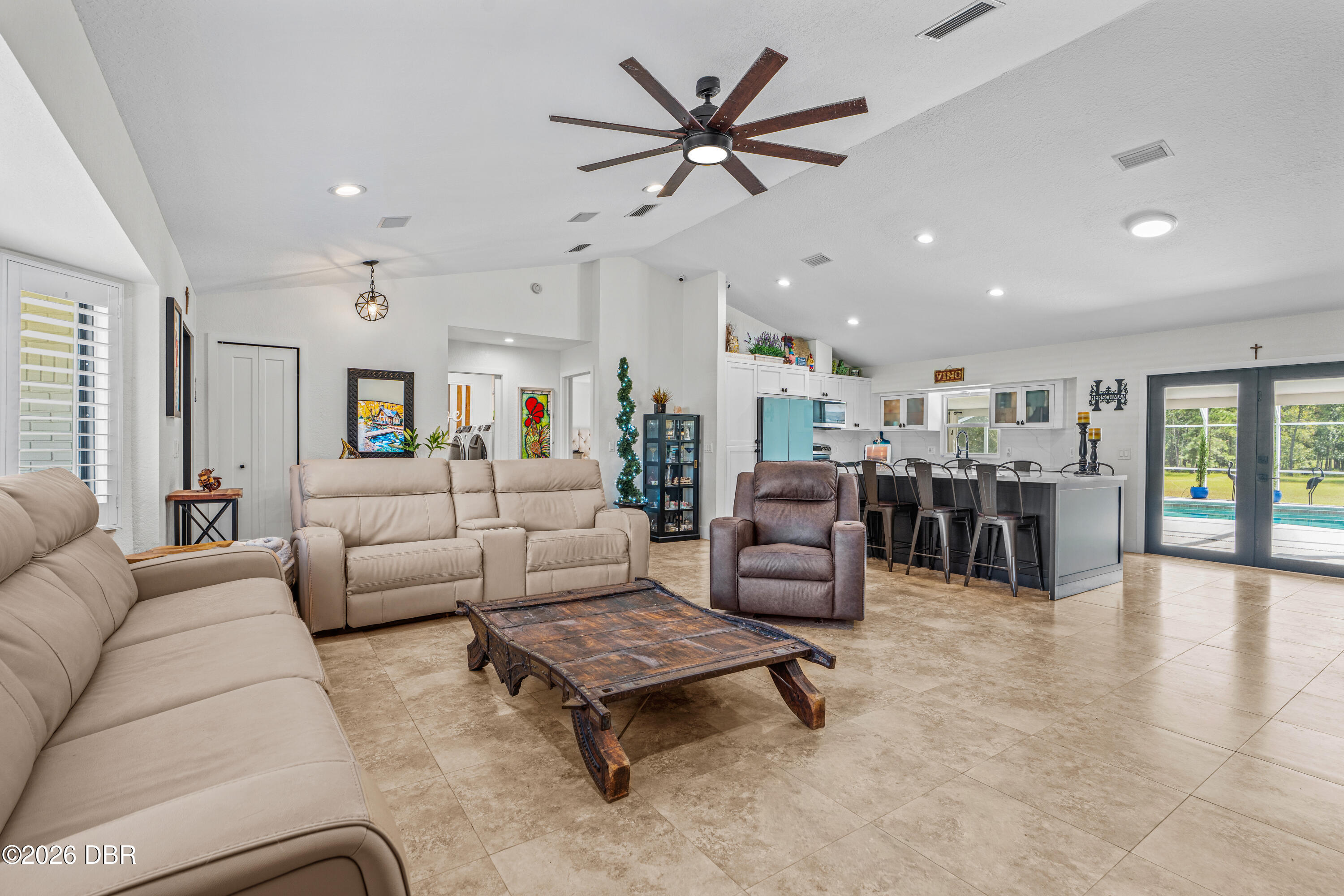 Open-plan living room and kitchen with beige sofas, a rustic coffee table, and a pool visible through sliding doors.
