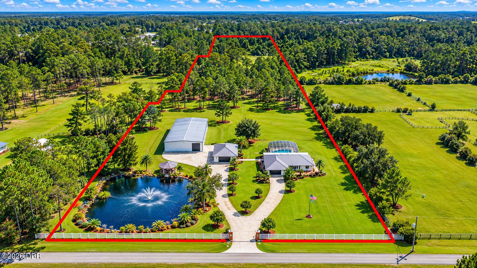 Aerial view of a landscaped property with a pond, fountain, home, outbuildings, and expansive lawns.