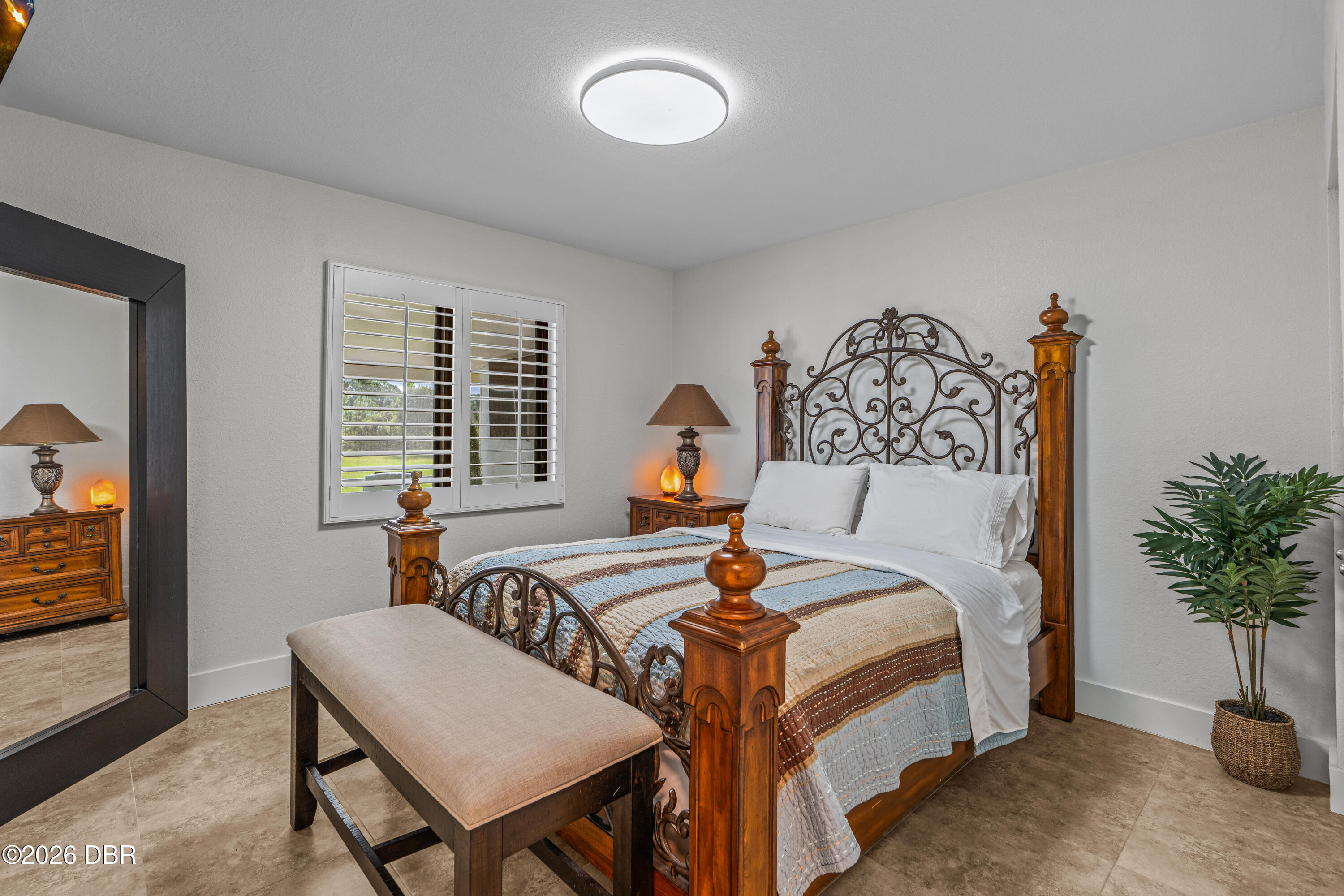 Staged bedroom with an ornate four-poster bed, plantation shutters, and a cushioned bench.