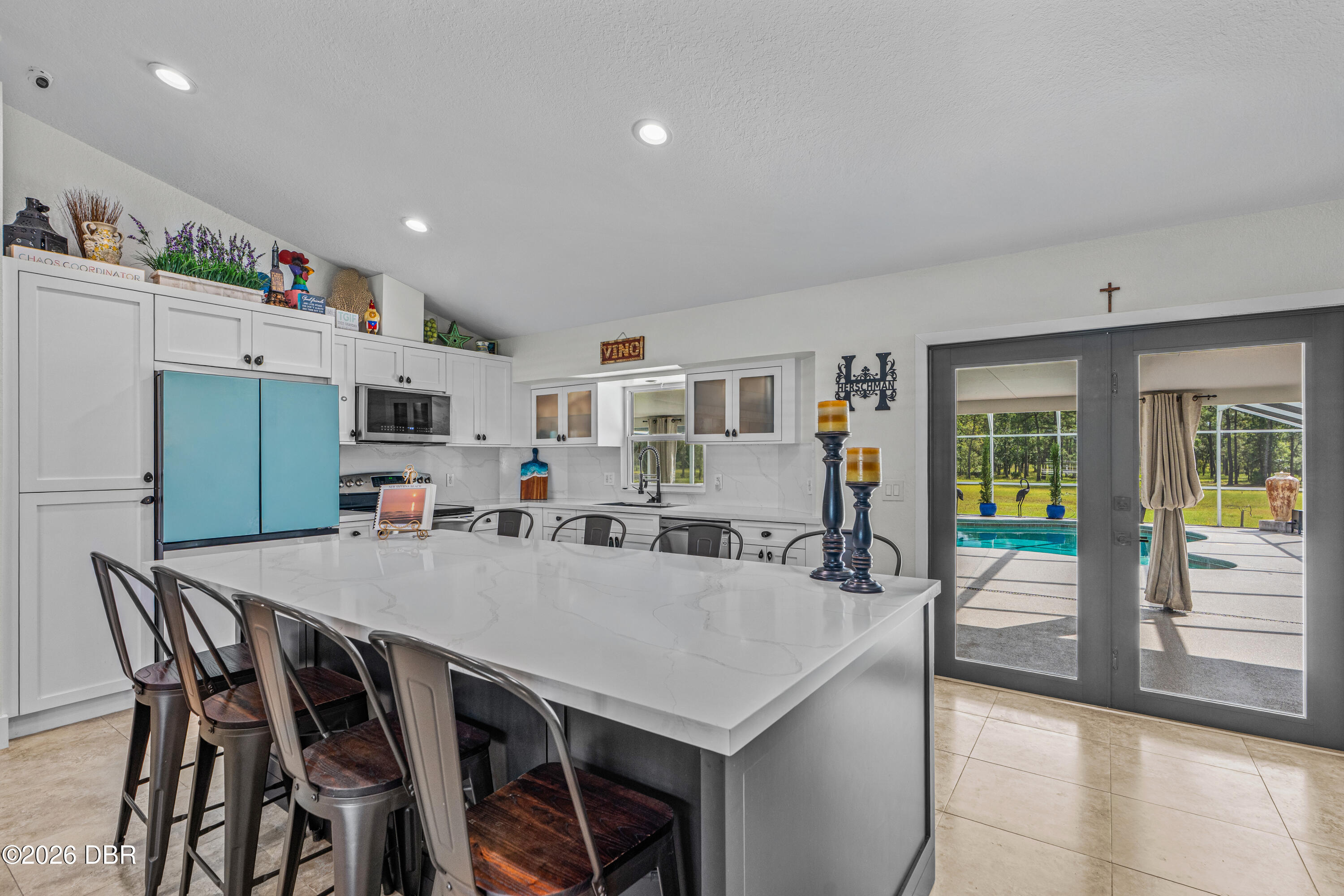 Kitchen with a large white island and blue refrigerator, opening through glass doors to a pool.
