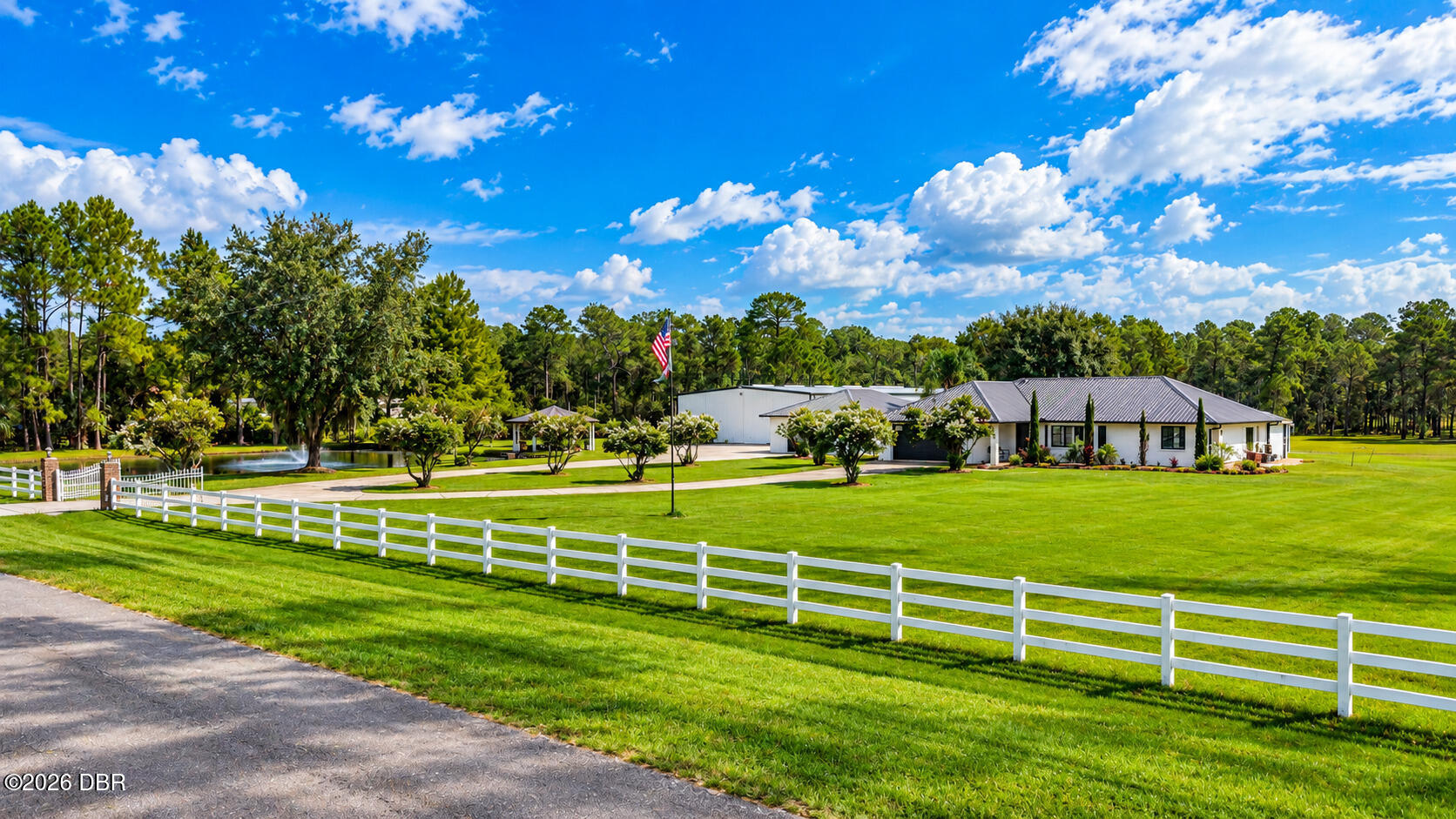 Low white building with expansive lawns, a white fence, and a pond with fountain.