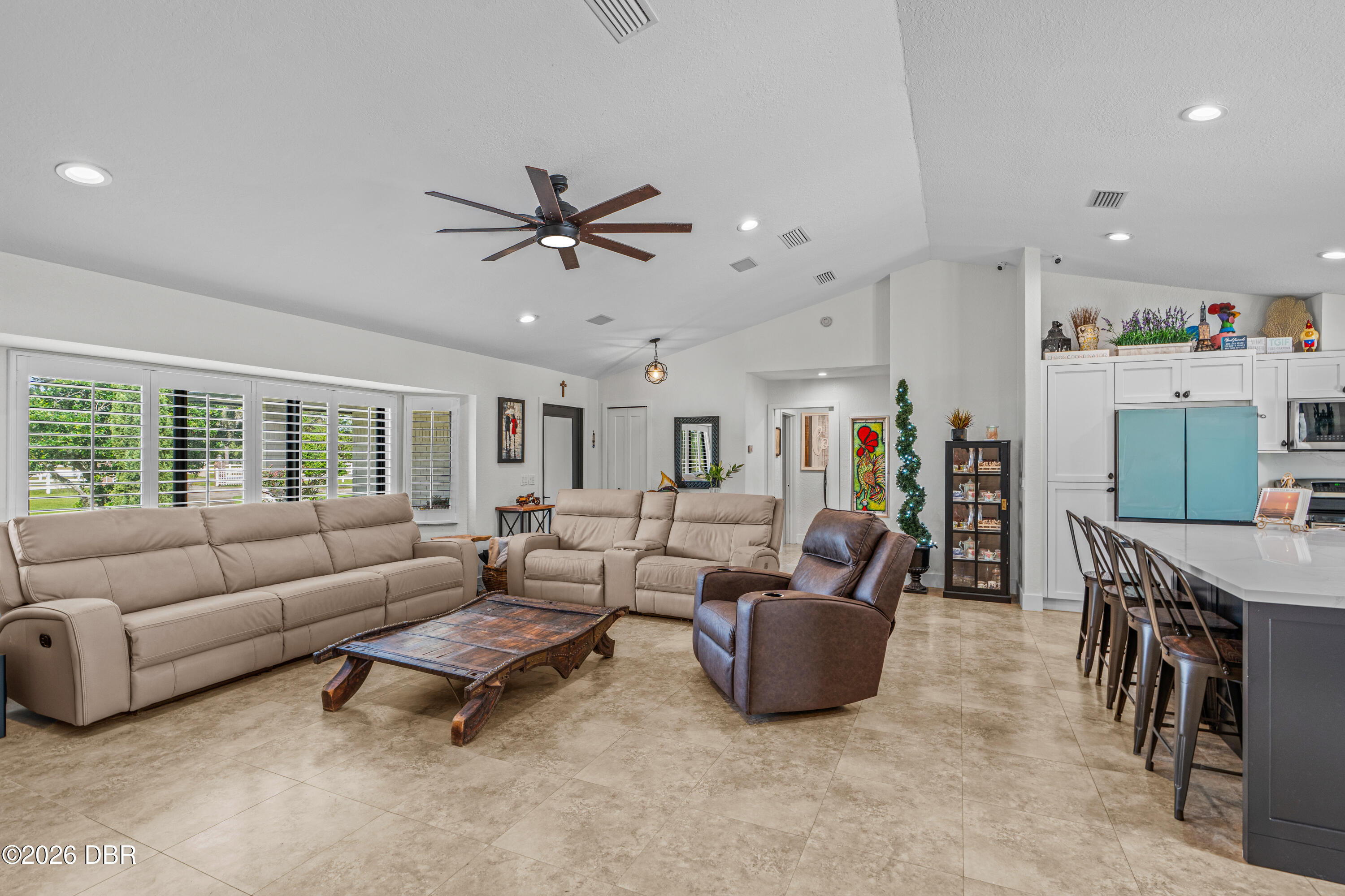 Open-plan living room with beige reclining sofas, a rustic coffee table, and a kitchen island with bar stools.