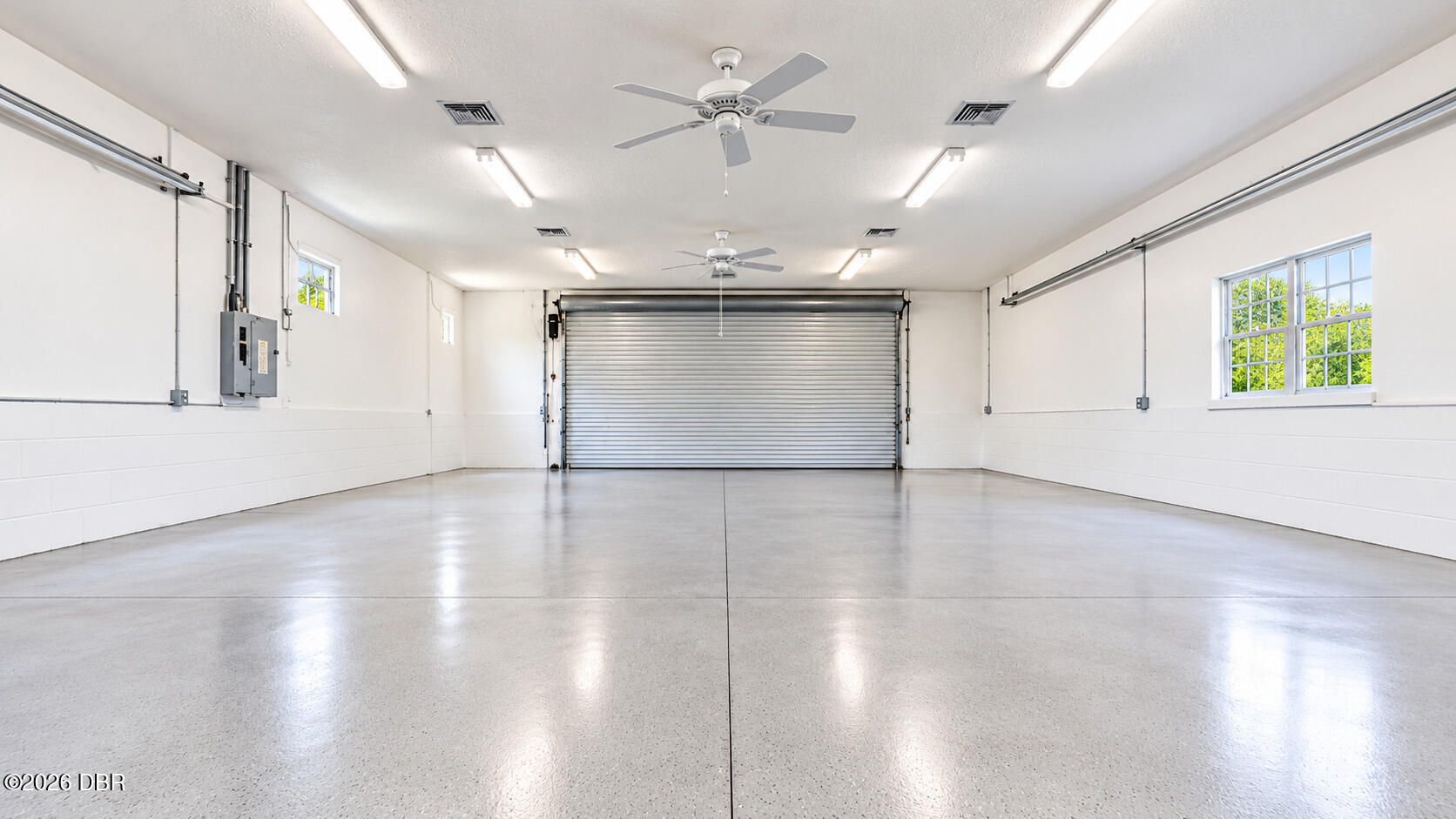 Empty garage with a wide roll-up door, smooth concrete floor, ceiling fans, and windows.