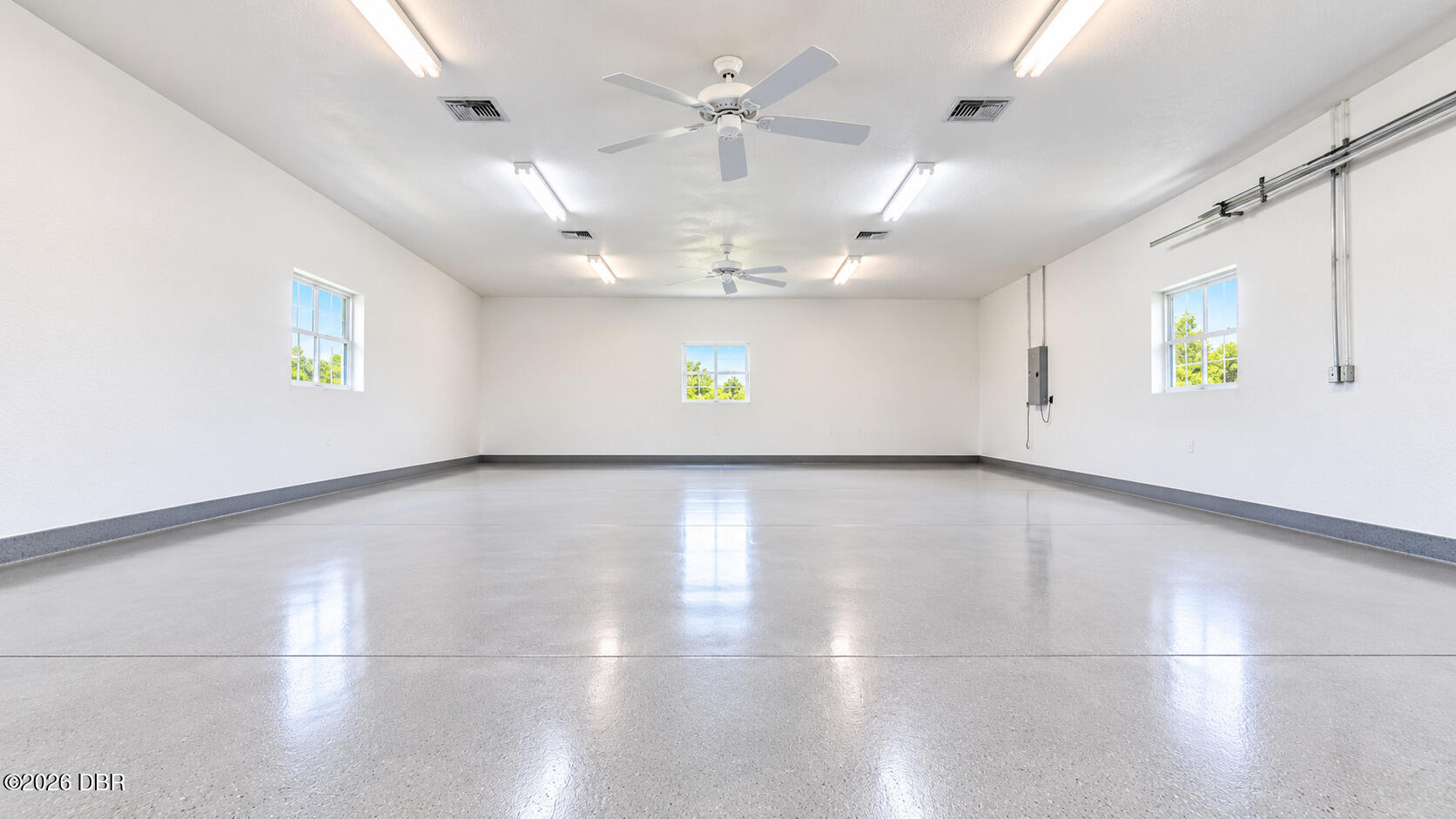 Empty garage with a smooth floor, three windows, ceiling fans, and fluorescent lighting.
