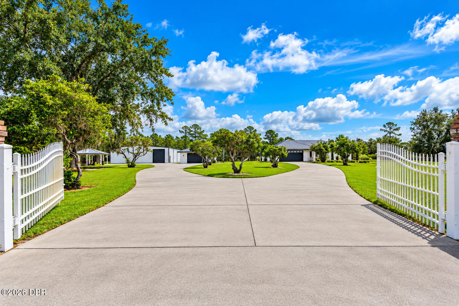 Property entrance with open white gates and a curved driveway leading to a modern building.