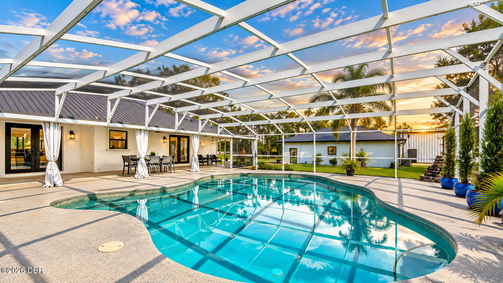 Screened pool patio with curved swimming pool, white pergola, and outdoor dining area at sunset.