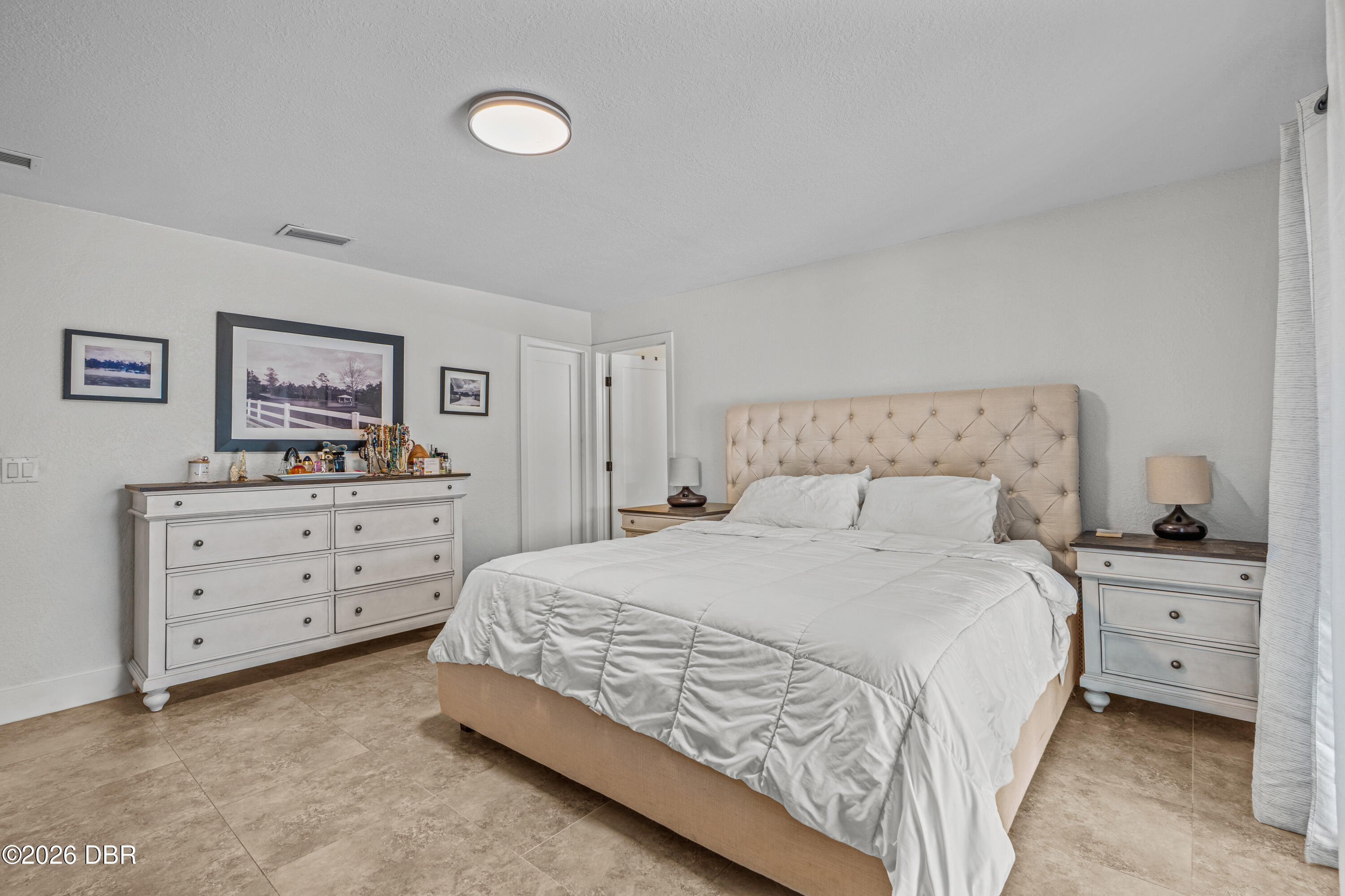 Bedroom with a tufted beige headboard, white bedding, matching dressers, and tile flooring.