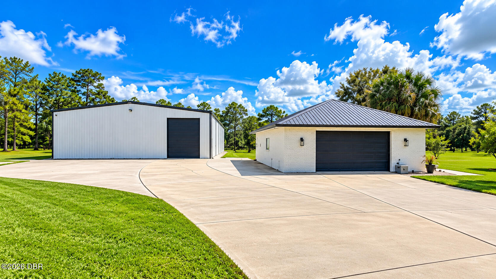 White metal outbuilding and separate brick garage beside a broad concrete driveway.