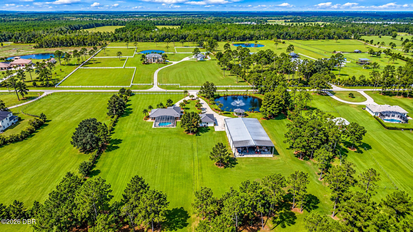 Aerial view of a sprawling green property with a home, pool, fenced paddocks, and a pond with a fountain.