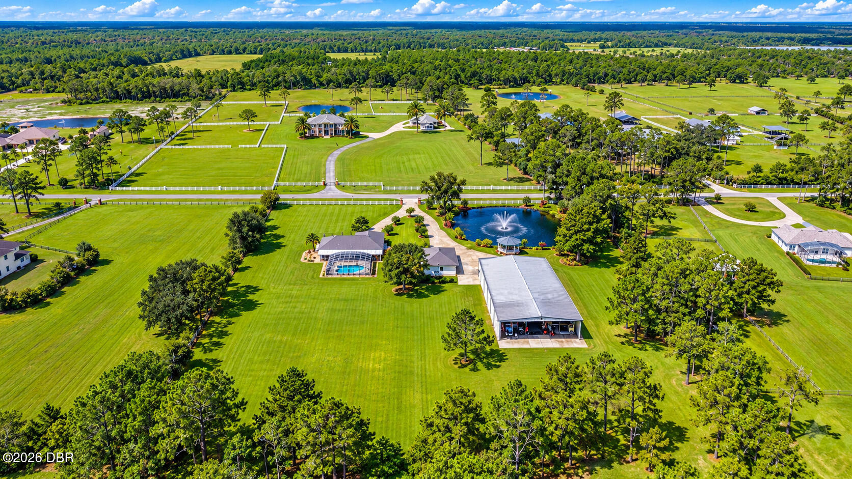 Aerial view of a sprawling estate with fenced paddocks, a pool, fountain pond, and wooded grounds.