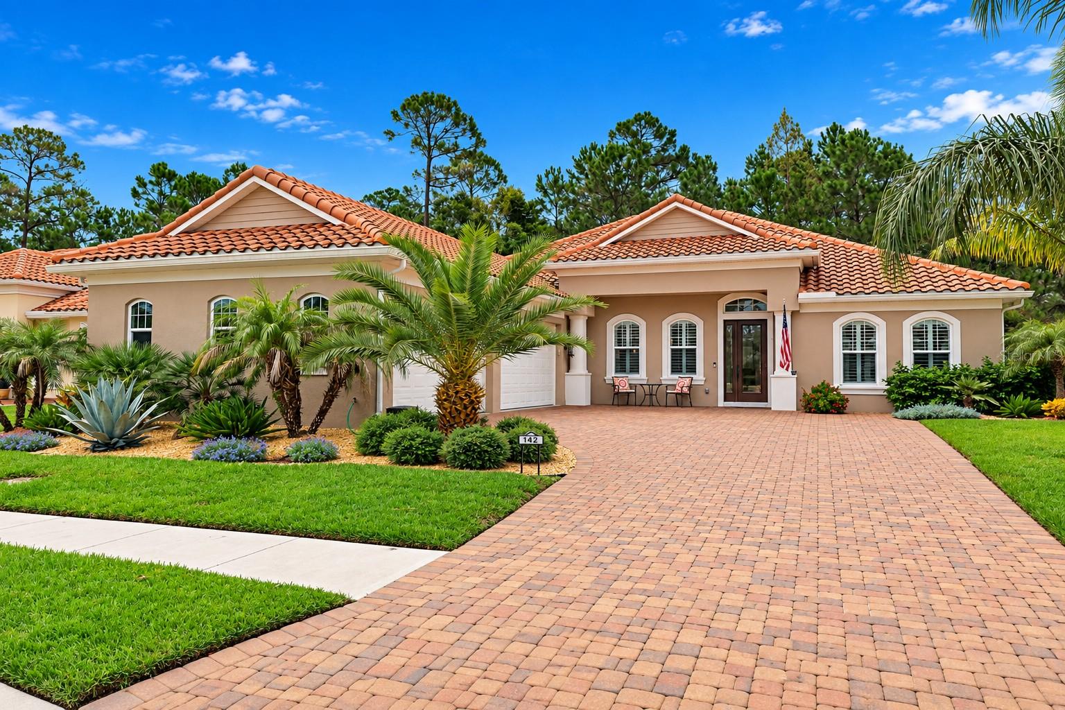 Single-story stucco home with a terracotta tile roof, palm landscaping, and a wide brick-paver driveway.