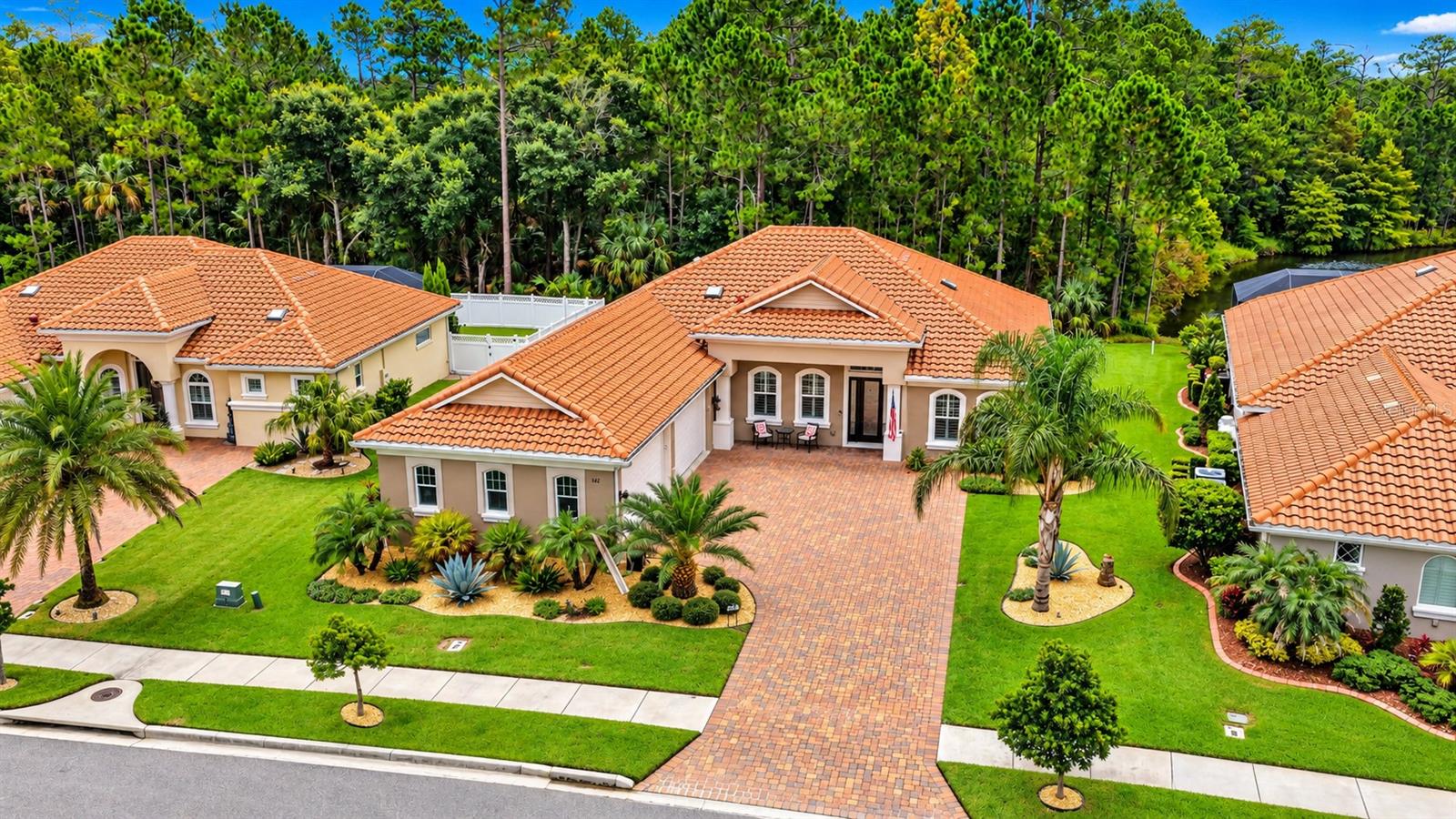 Aerial view of a beige home with a terracotta tile roof, brick-paver driveway, and tropical landscaping.