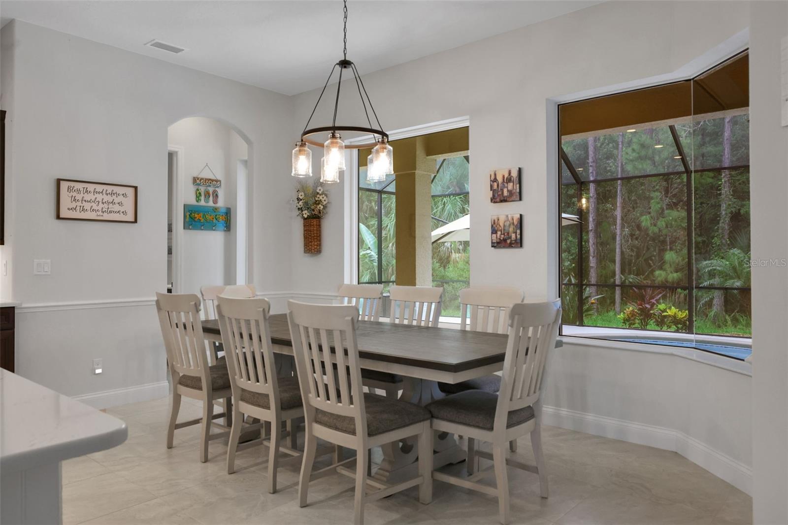 Dining room with a white table and chairs beside large windows overlooking a screened patio and greenery.