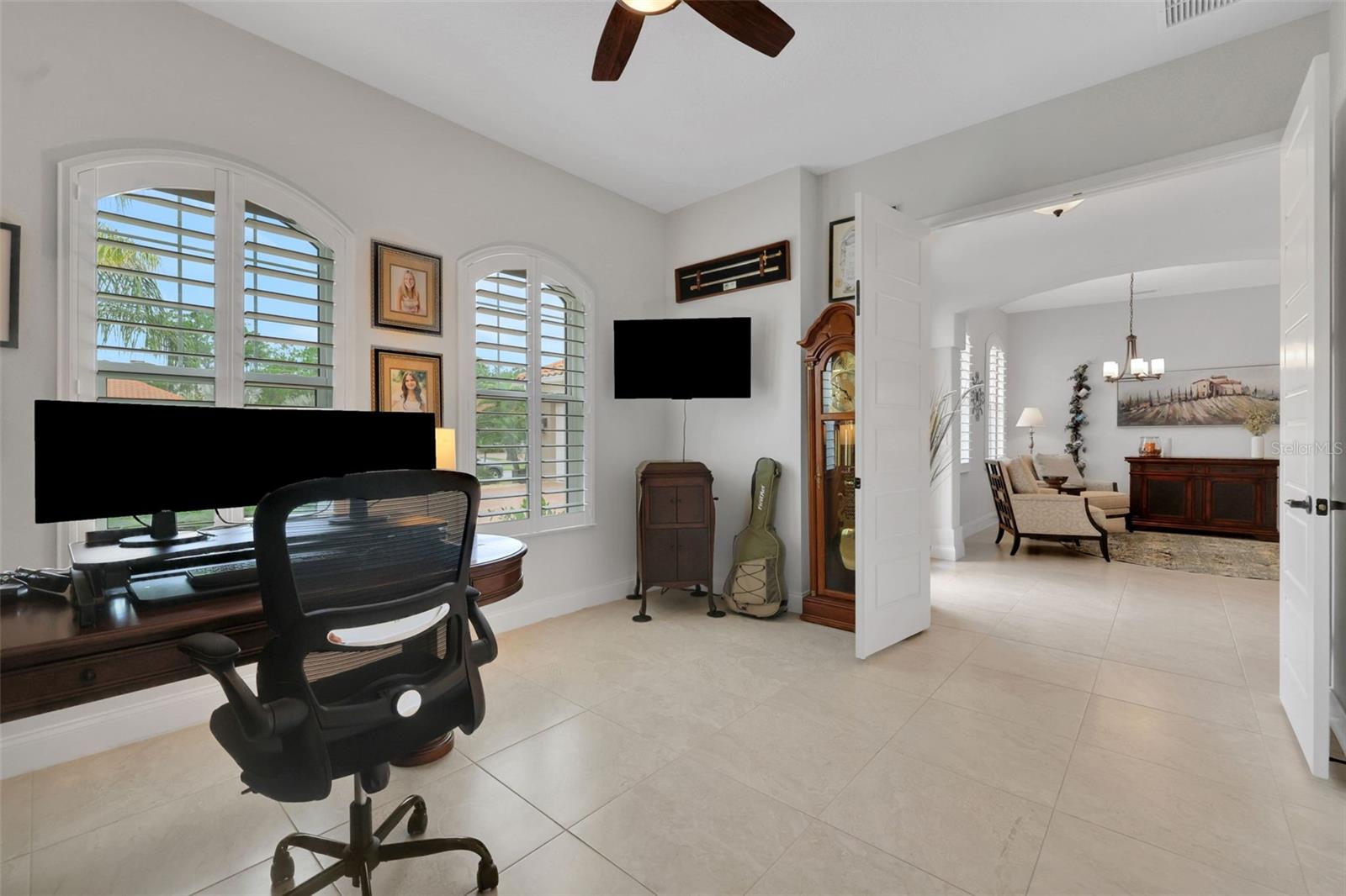 Home office with arched shuttered windows, dark wood desk, and open doors to a sitting area.