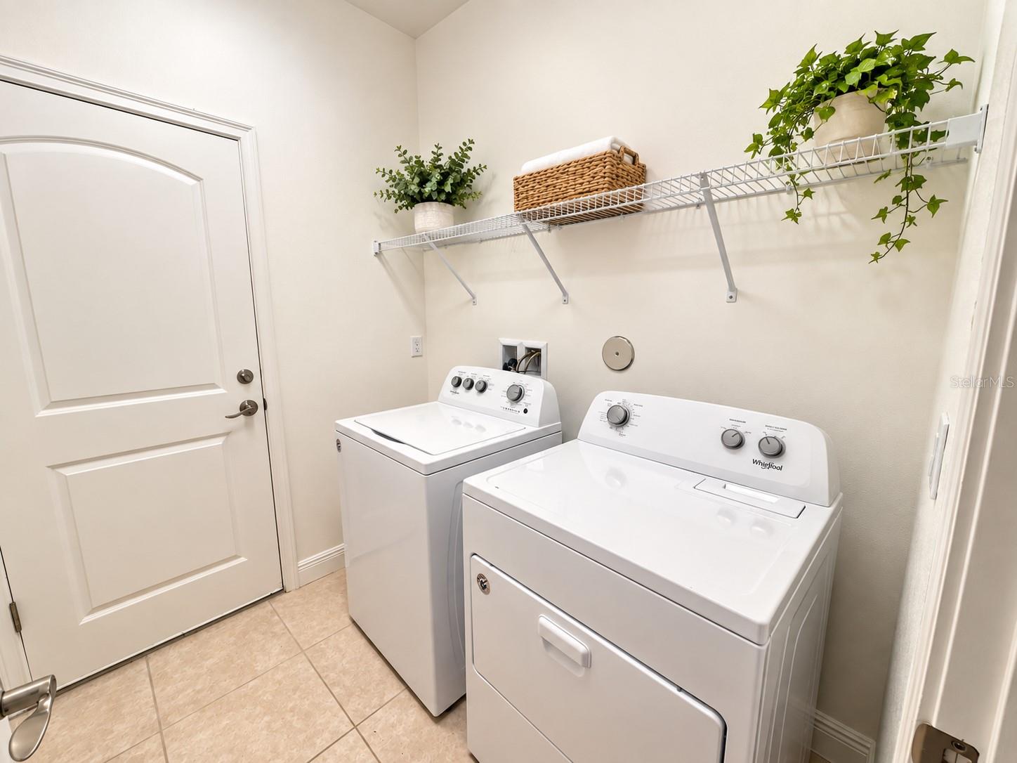 Laundry room with white washer and dryer beneath a wire shelf decorated with plants.