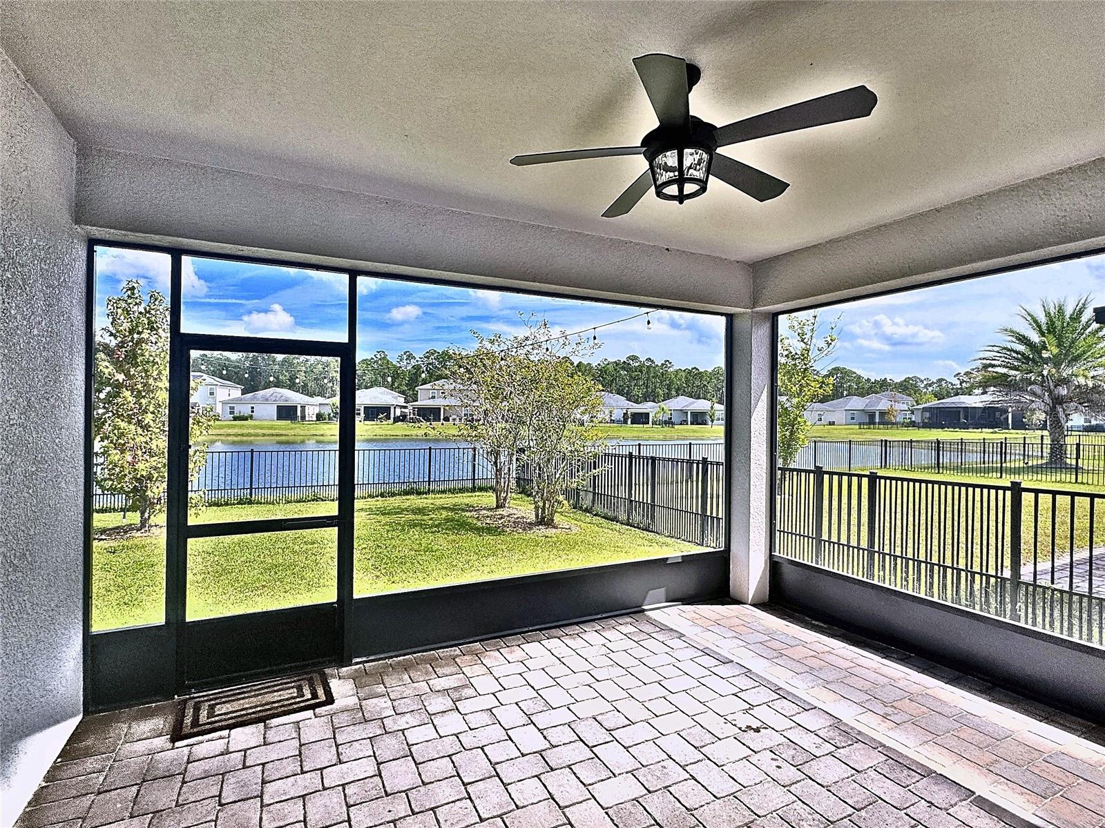 Screened lanai with paver flooring and ceiling fan overlooking a pond and fenced lawn.