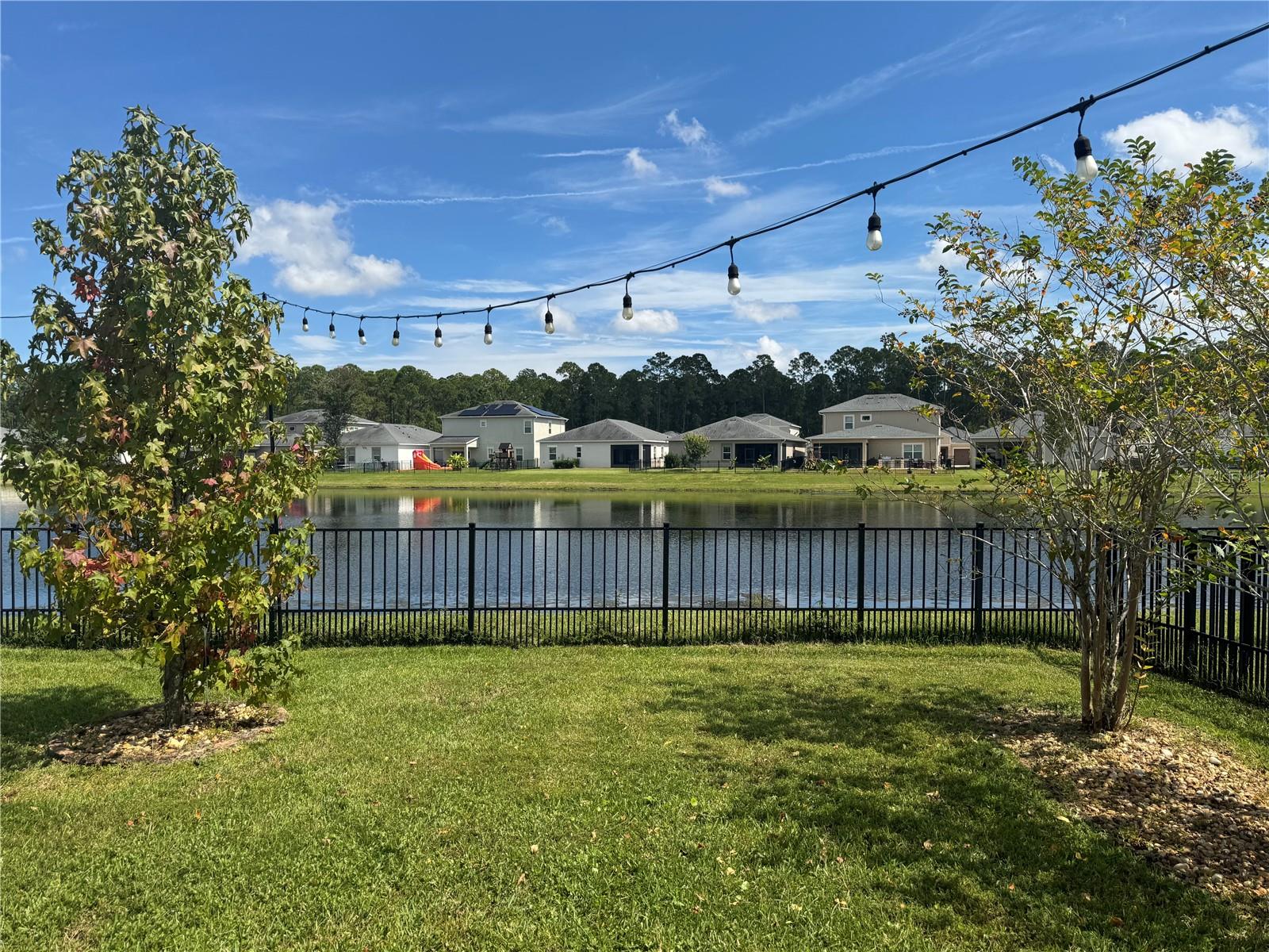 Backyard overlooking a neighborhood lake, framed by lawn, trees, and overhead string lights.