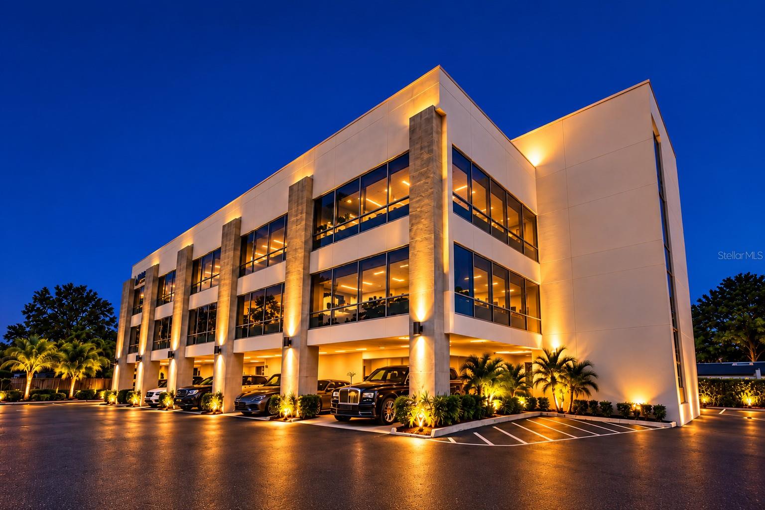 Modern office building illuminated at dusk, with glass windows, palms, and a landscaped parking area.