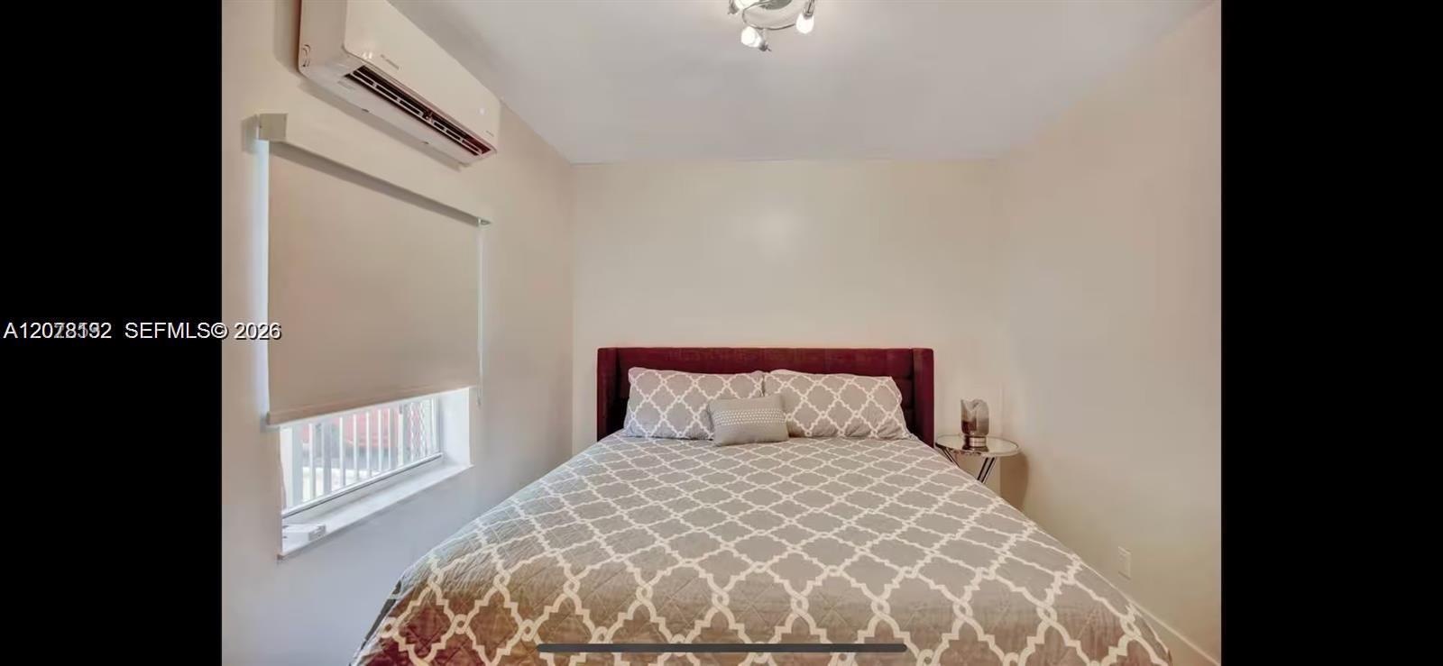Bedroom with a patterned bedspread, burgundy headboard, shaded window, and wall-mounted air conditioner.