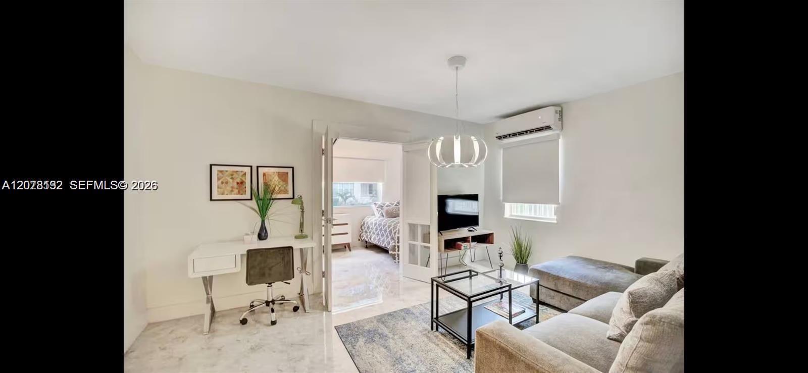 Furnished apartment living room with a gray sectional, glass coffee table, and desk beside an open bedroom doorway.