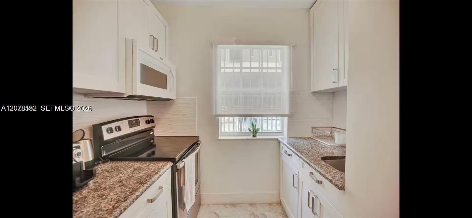 Bright narrow kitchen with white cabinets, granite counters, stainless appliances, and a shaded window.