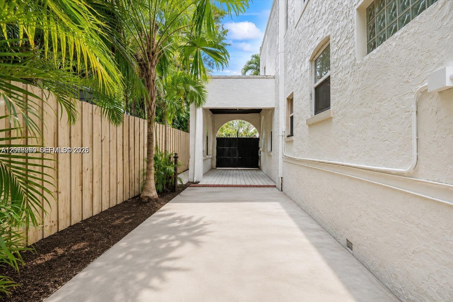 Paved driveway beside a white stucco building, leading to an arched entry and black gate among tropical palms.