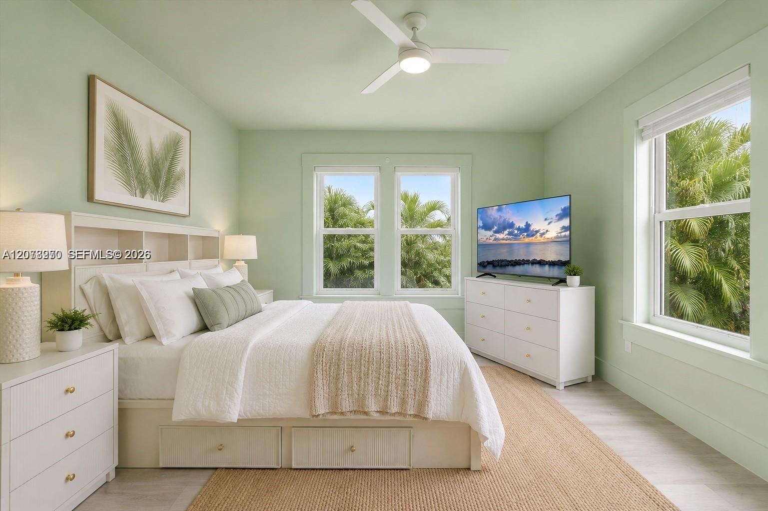 Bedroom with a white bed, pale green walls, and windows overlooking tropical palms.