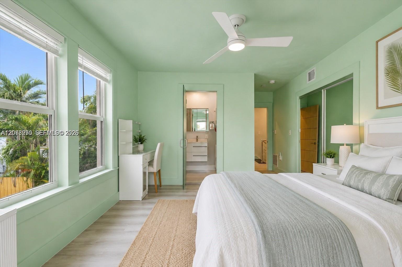 Mint-green bedroom with a white bed, large windows, and a bathroom doorway.