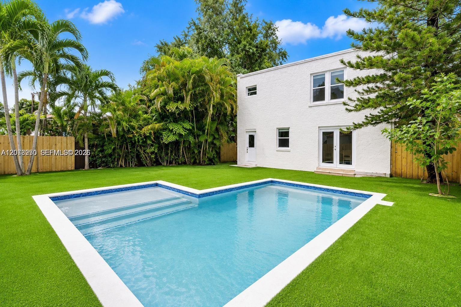 Backyard swimming pool beside a white two-story house, surrounded by lawn and tropical landscaping.