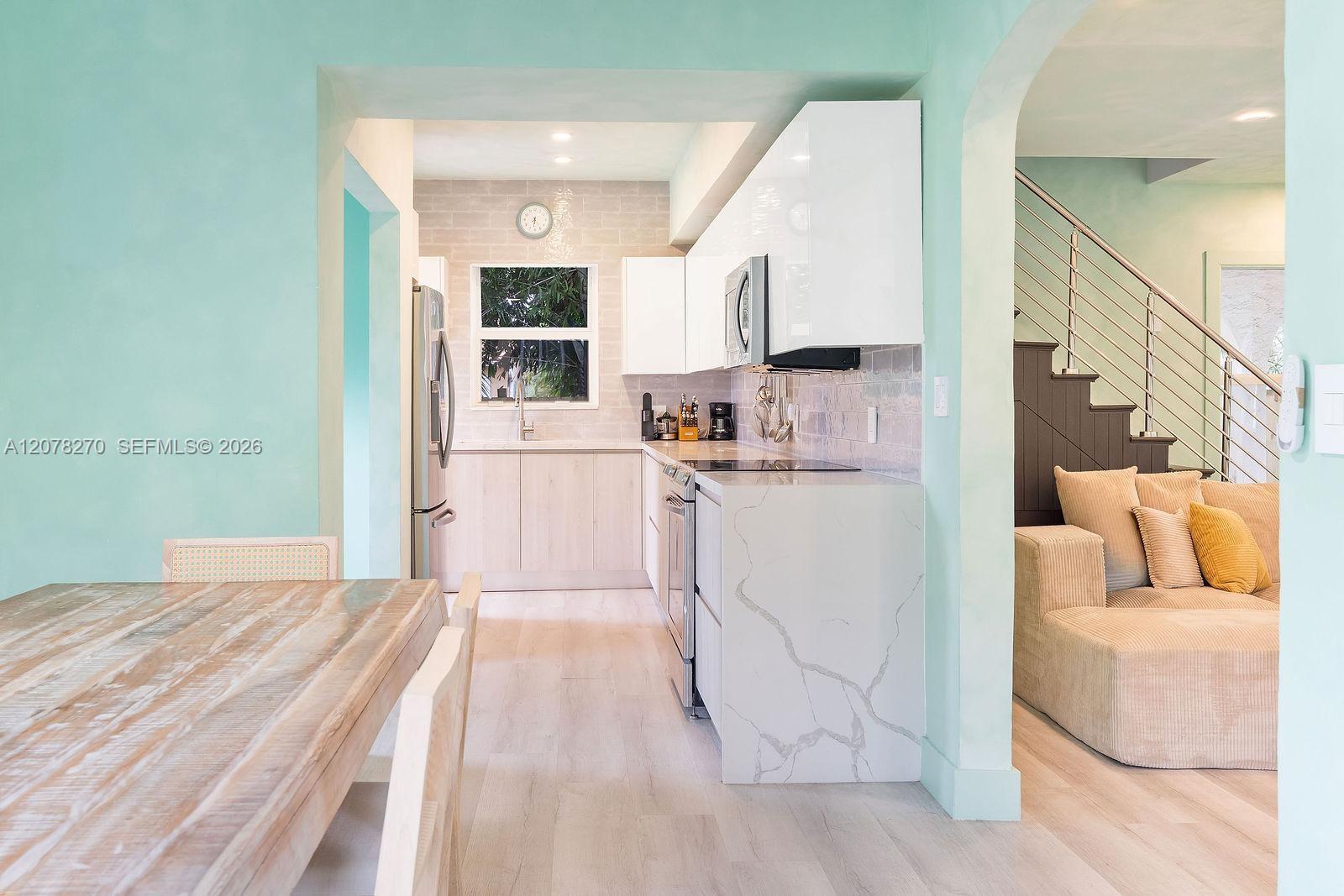 Bright white kitchen with marble-look island, opening to a living area with beige sectional and staircase.