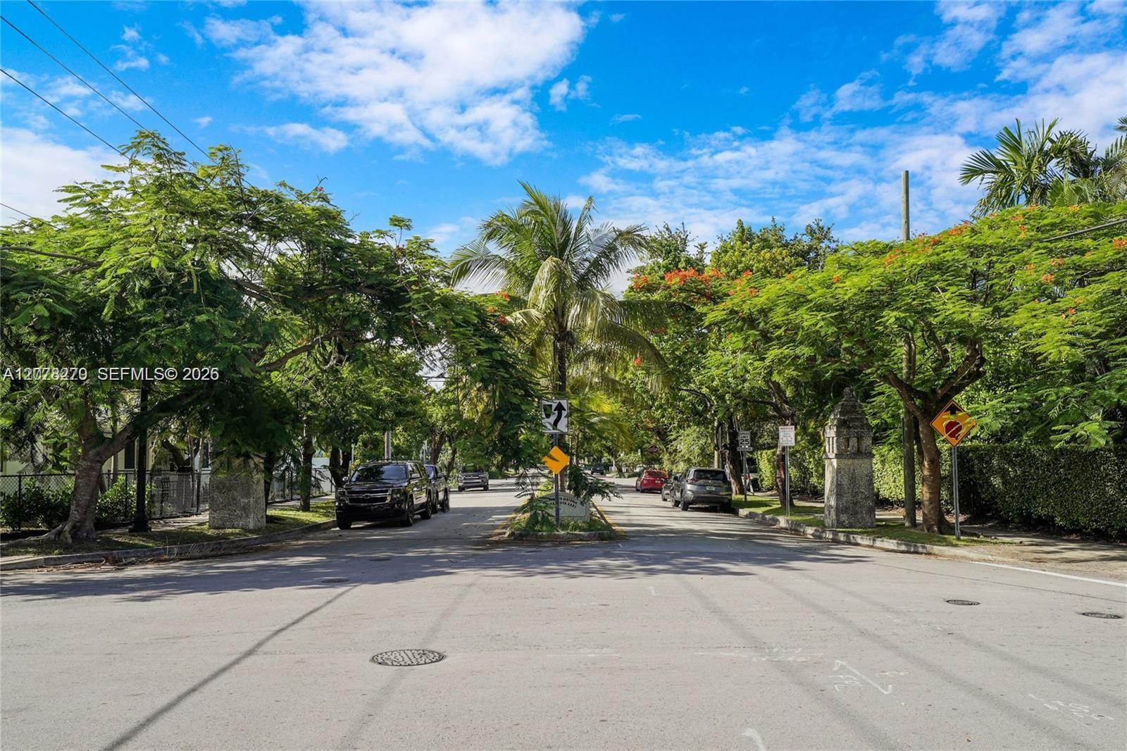 Tree-lined residential street with palm trees, parked cars, and stone gateposts.