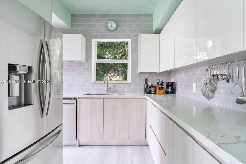 Bright modern kitchen with white cabinetry, gray tile backsplash, stainless steel refrigerator, and long counters.