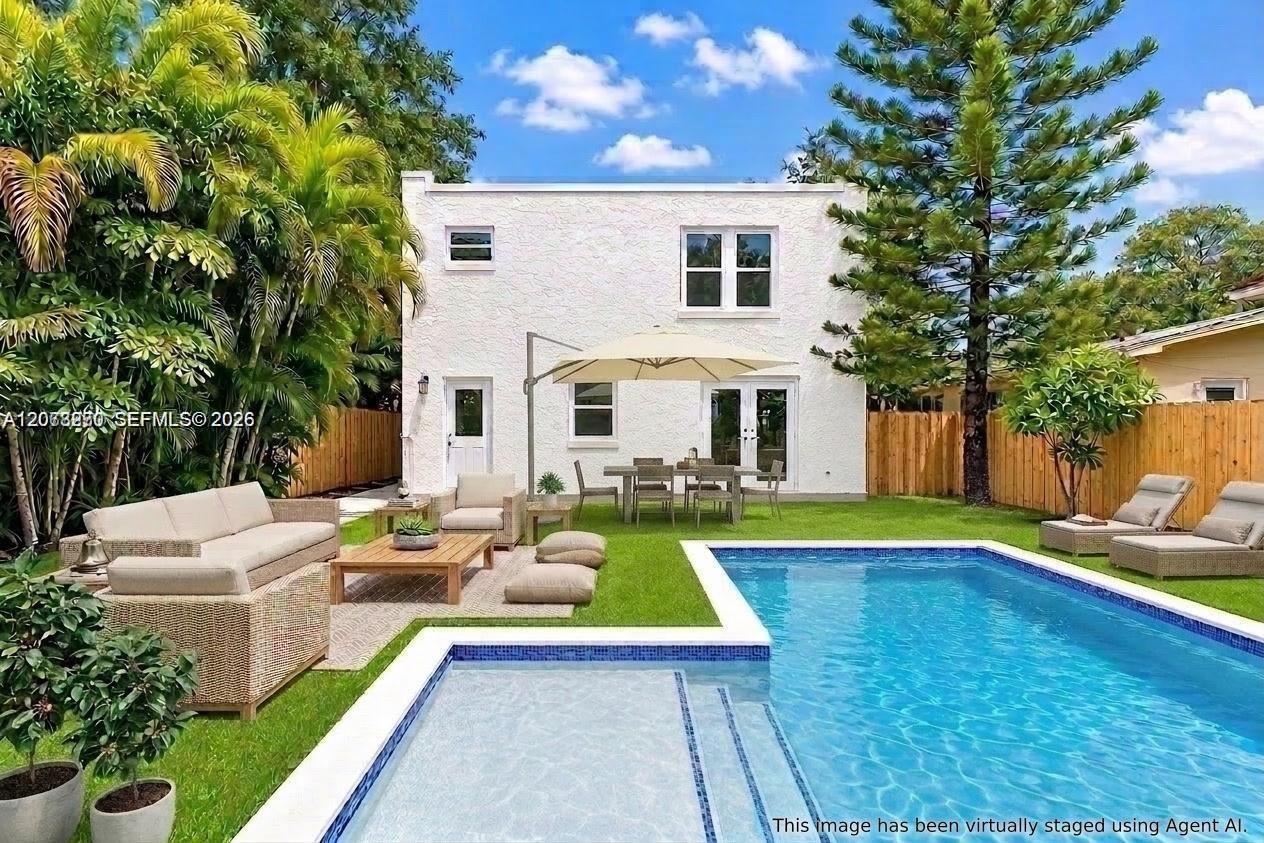White two-story house behind a blue pool and furnished patio, surrounded by tropical trees.