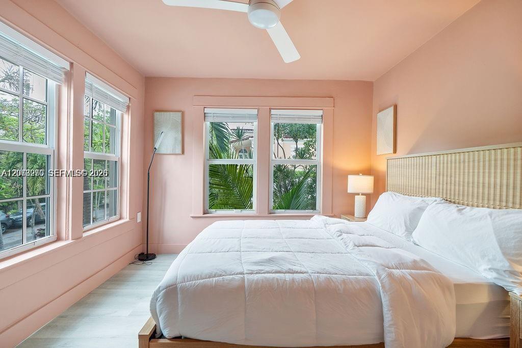 Bedroom with a white bed, peach-pink walls, and tall windows overlooking greenery.