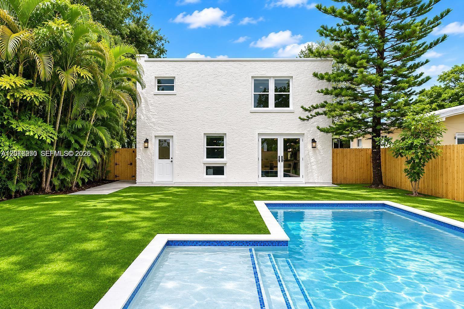 White stucco home behind a lawn and swimming pool, framed by tropical trees and wooden fencing.