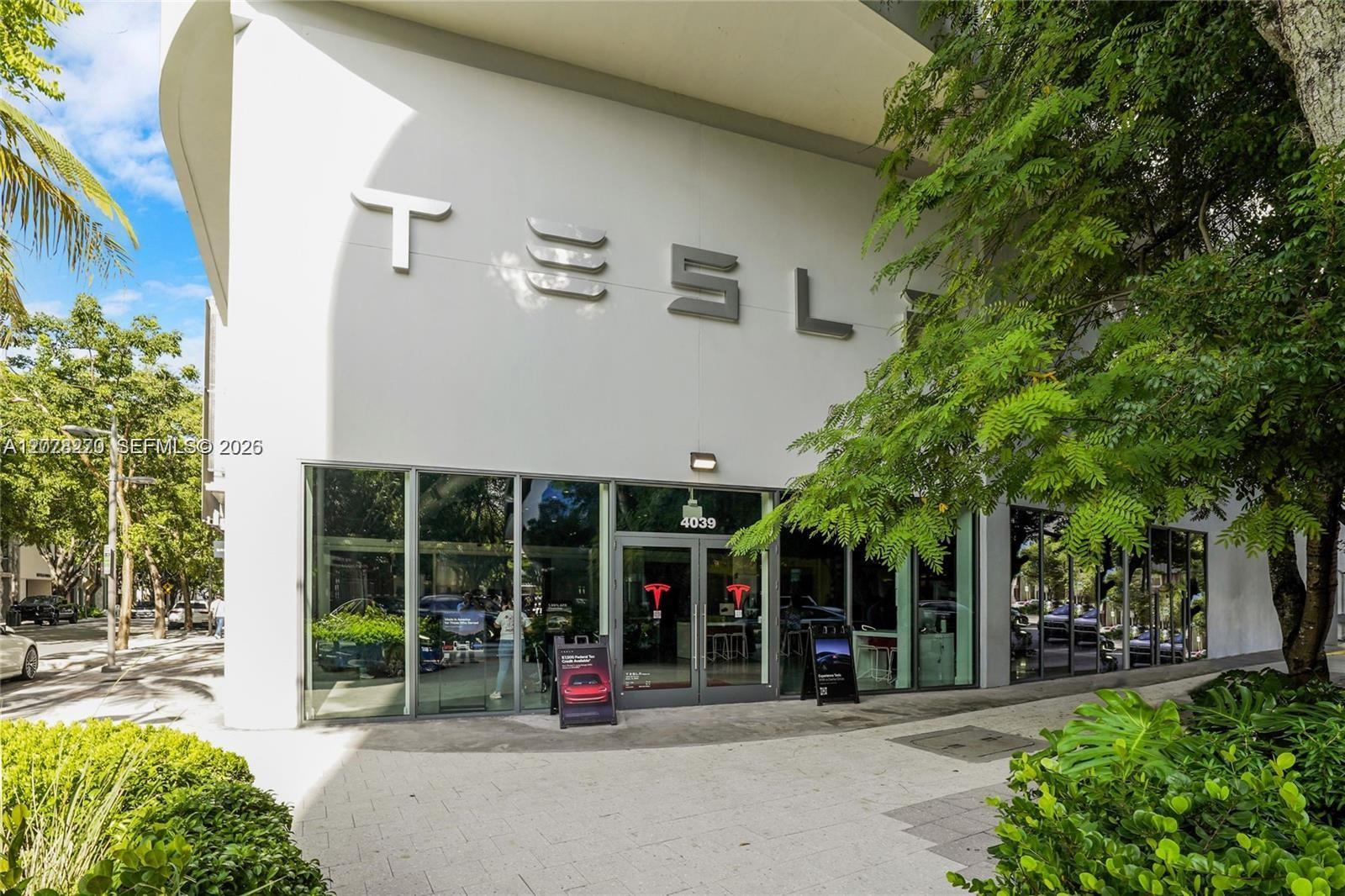 Tesla showroom exterior with a curved white facade, glass storefront, and leafy street trees.