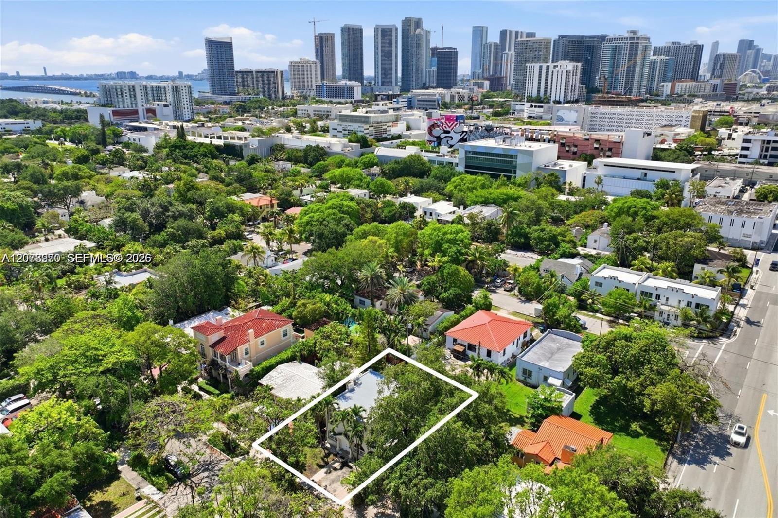 Aerial view of an outlined residential lot amid leafy homes, with Miami’s skyline and waterfront beyond.