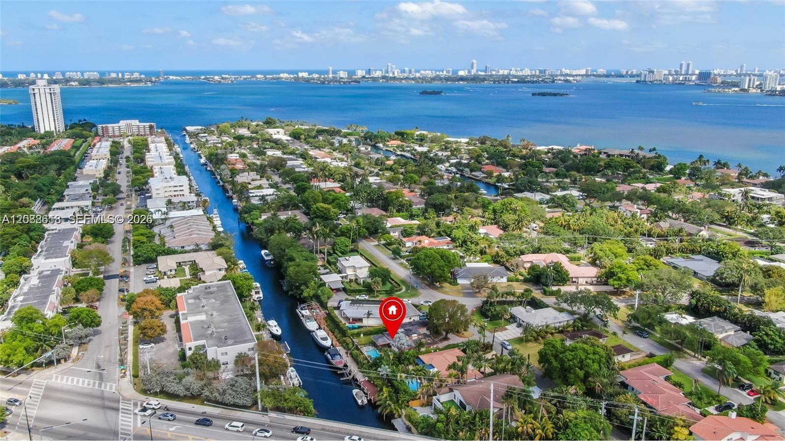 Aerial view of canal-front homes, Biscayne Bay, and the distant skyline, with a red pin marking a property.