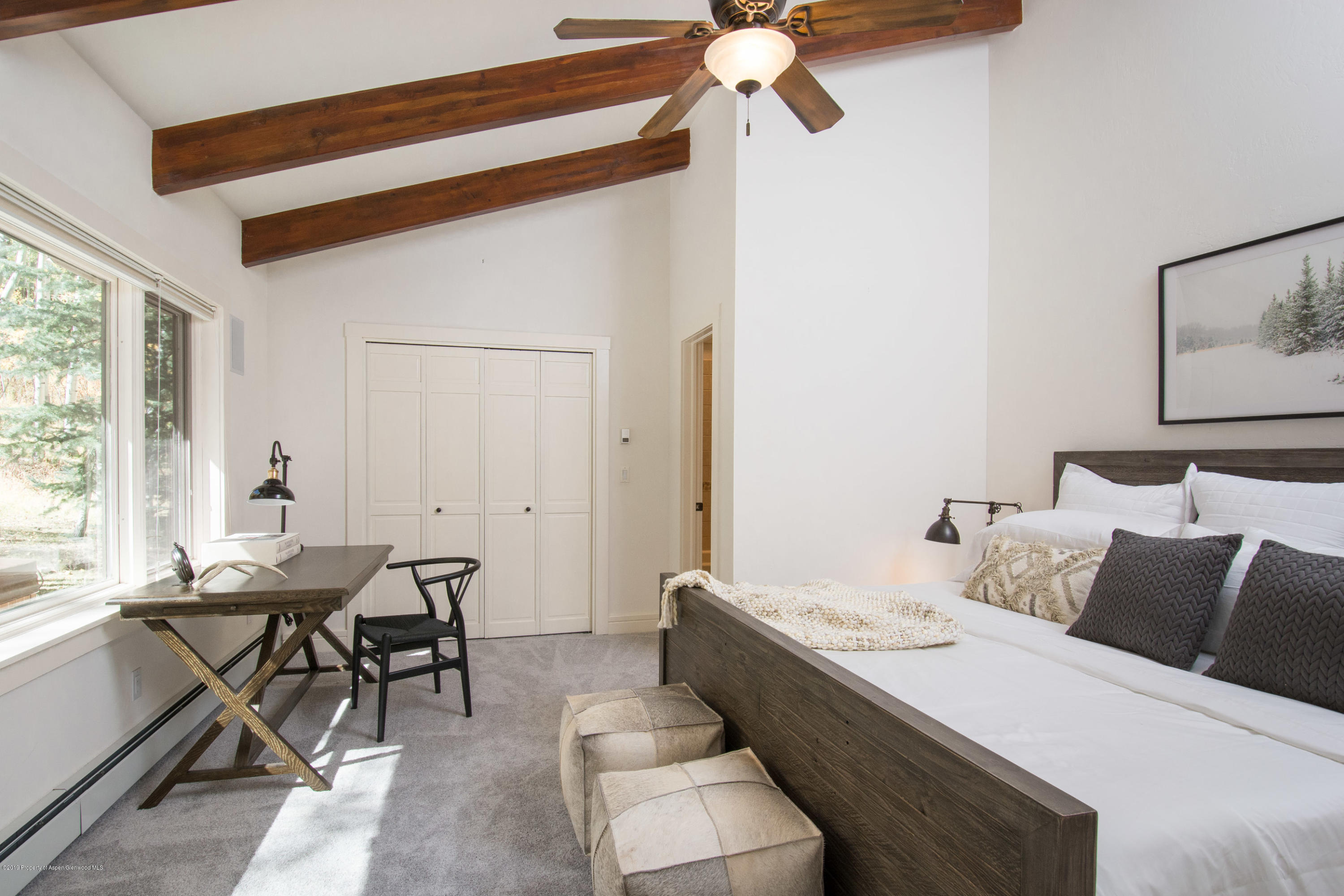 Bedroom with exposed wood ceiling beams, a dark wood bed, and a desk beside a large window.