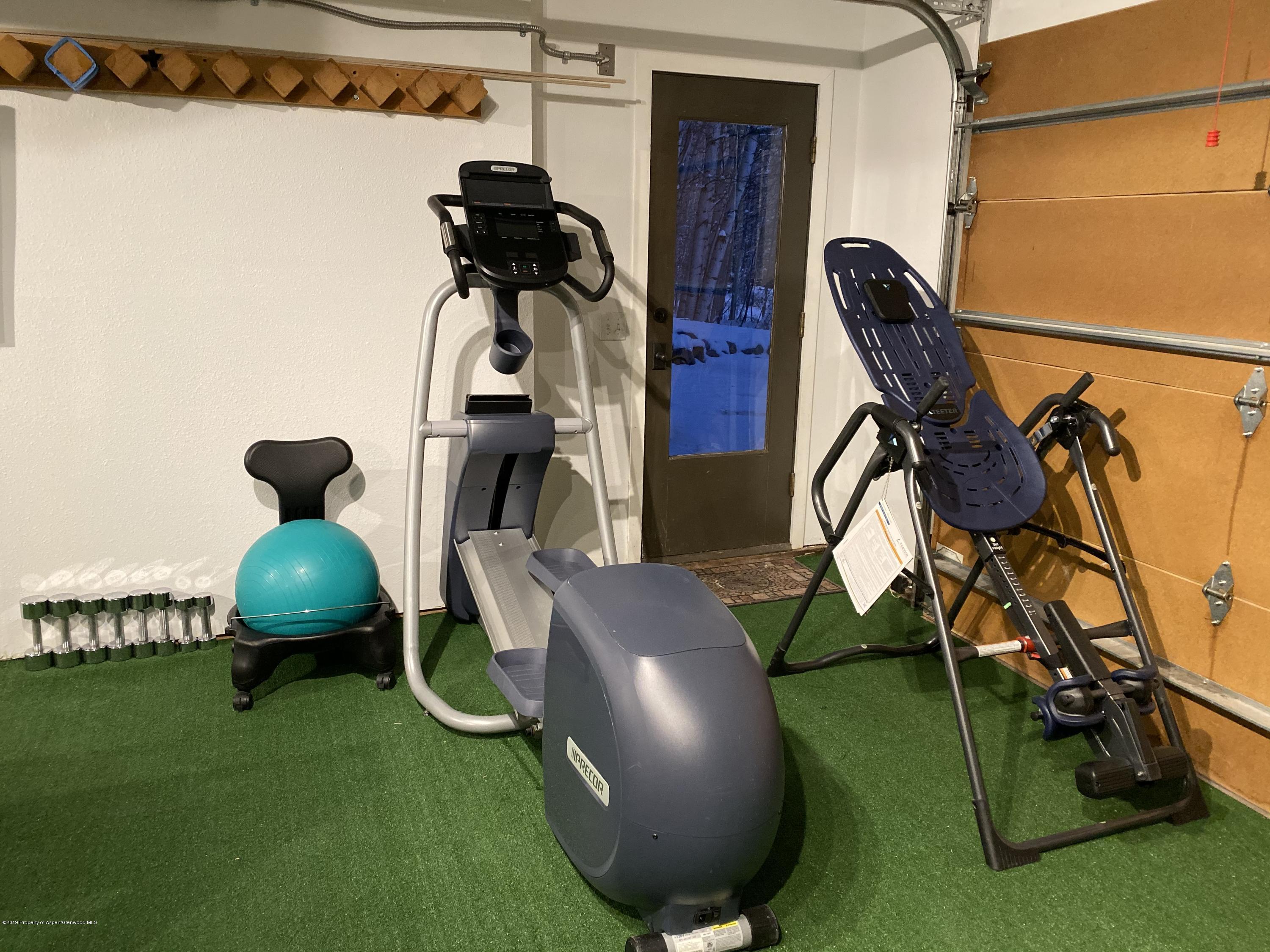 Home gym with an elliptical, inversion table, dumbbells, and turquoise exercise ball on green turf.