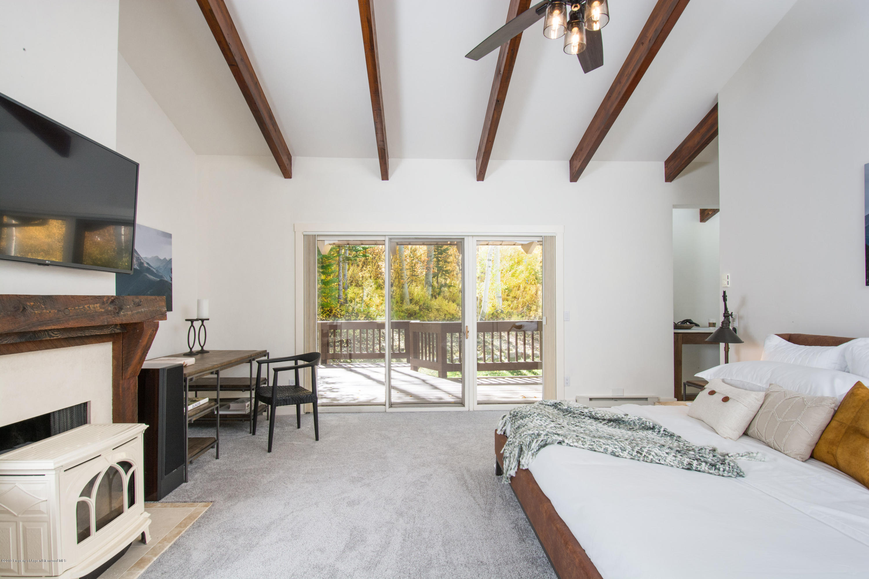 Bedroom with exposed wood beams, a fireplace, and sliding doors opening to a tree-lined deck.