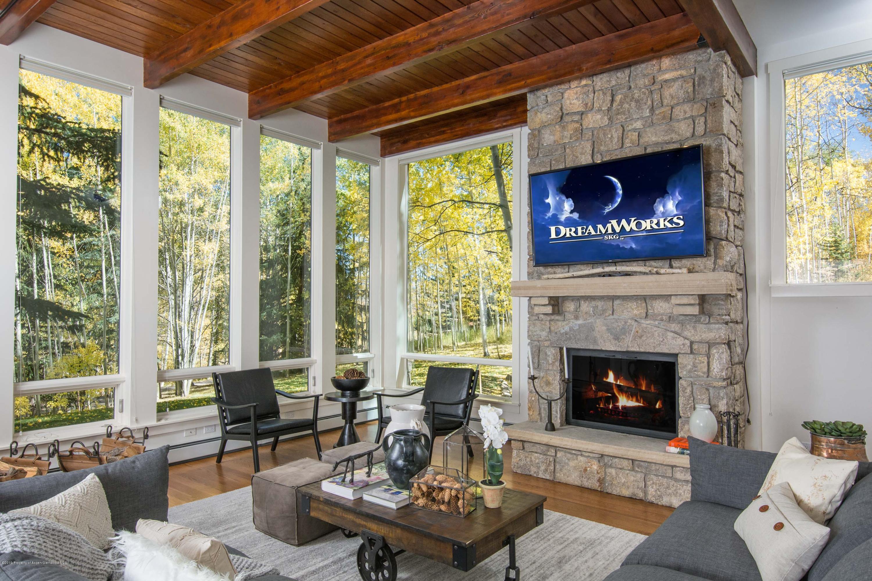 Furnished living room with a stone fireplace, exposed wood beams, and tall windows overlooking autumn trees.