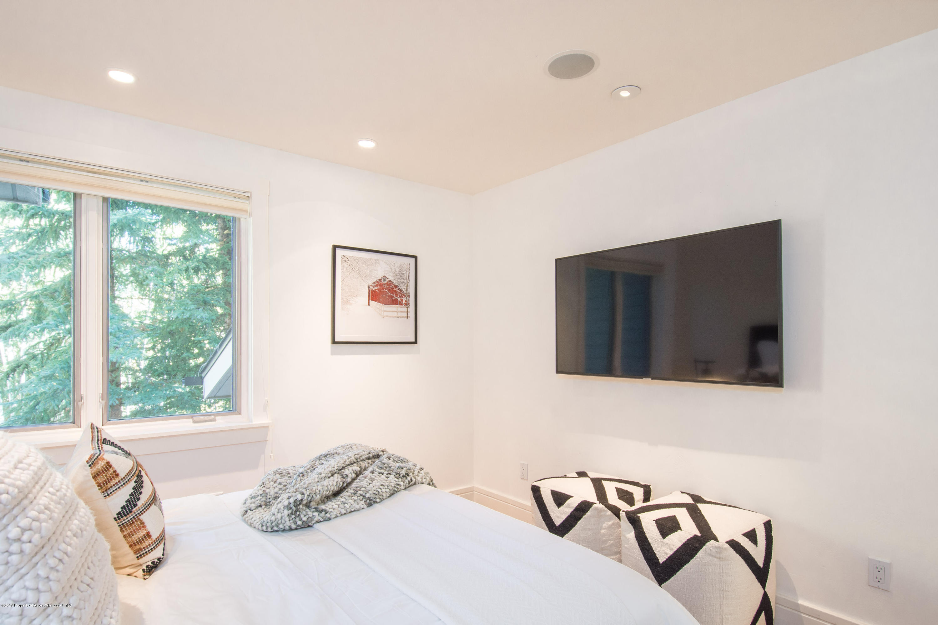 Furnished bedroom with a white bed, geometric stools, wall-mounted TV, and a window overlooking evergreen trees.