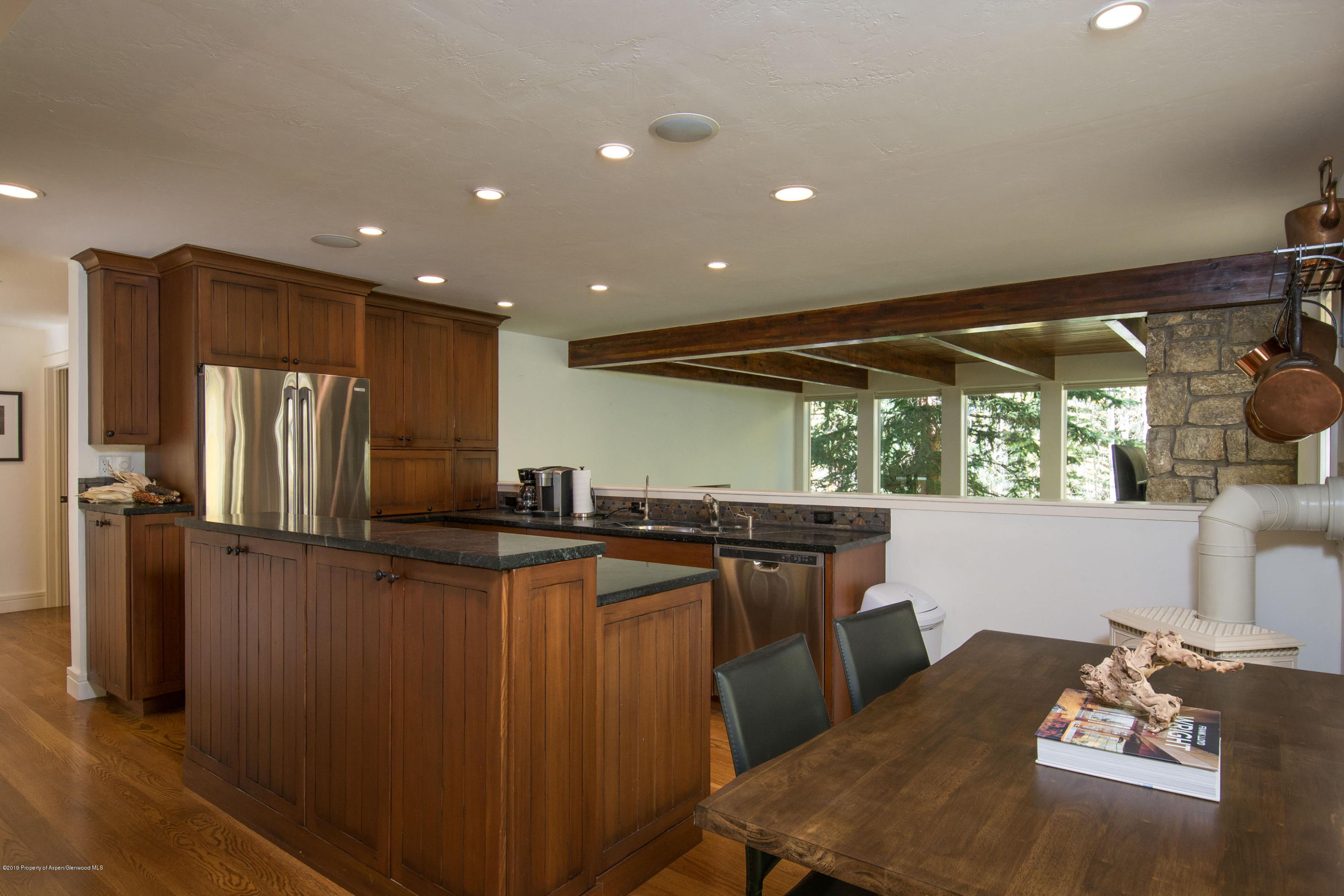 Kitchen and dining area with dark wood cabinets, stone counters, exposed beams, and windows overlooking trees.