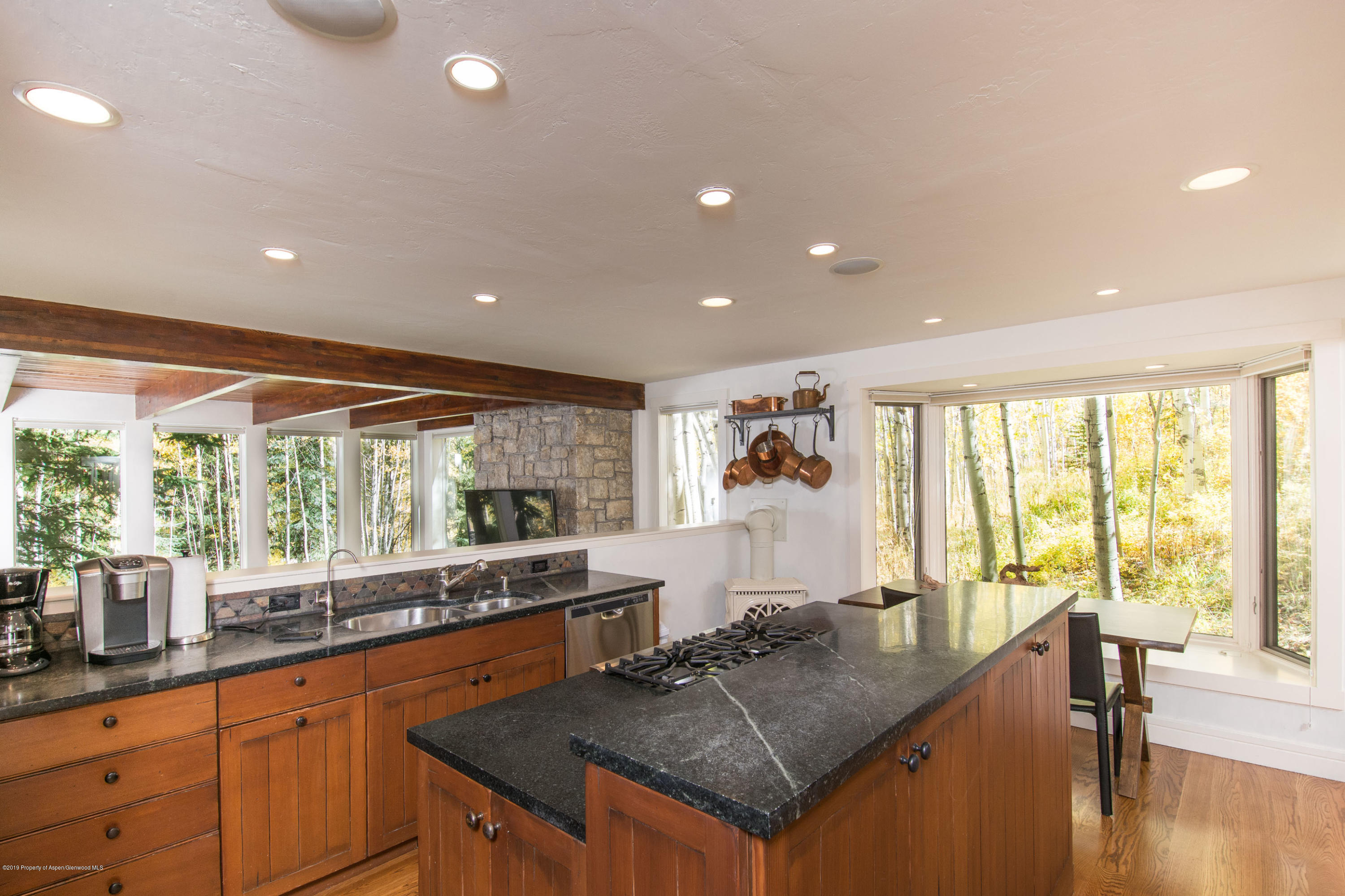 Kitchen with a large stone island, warm wood cabinets, exposed beams, and windows overlooking autumn trees.