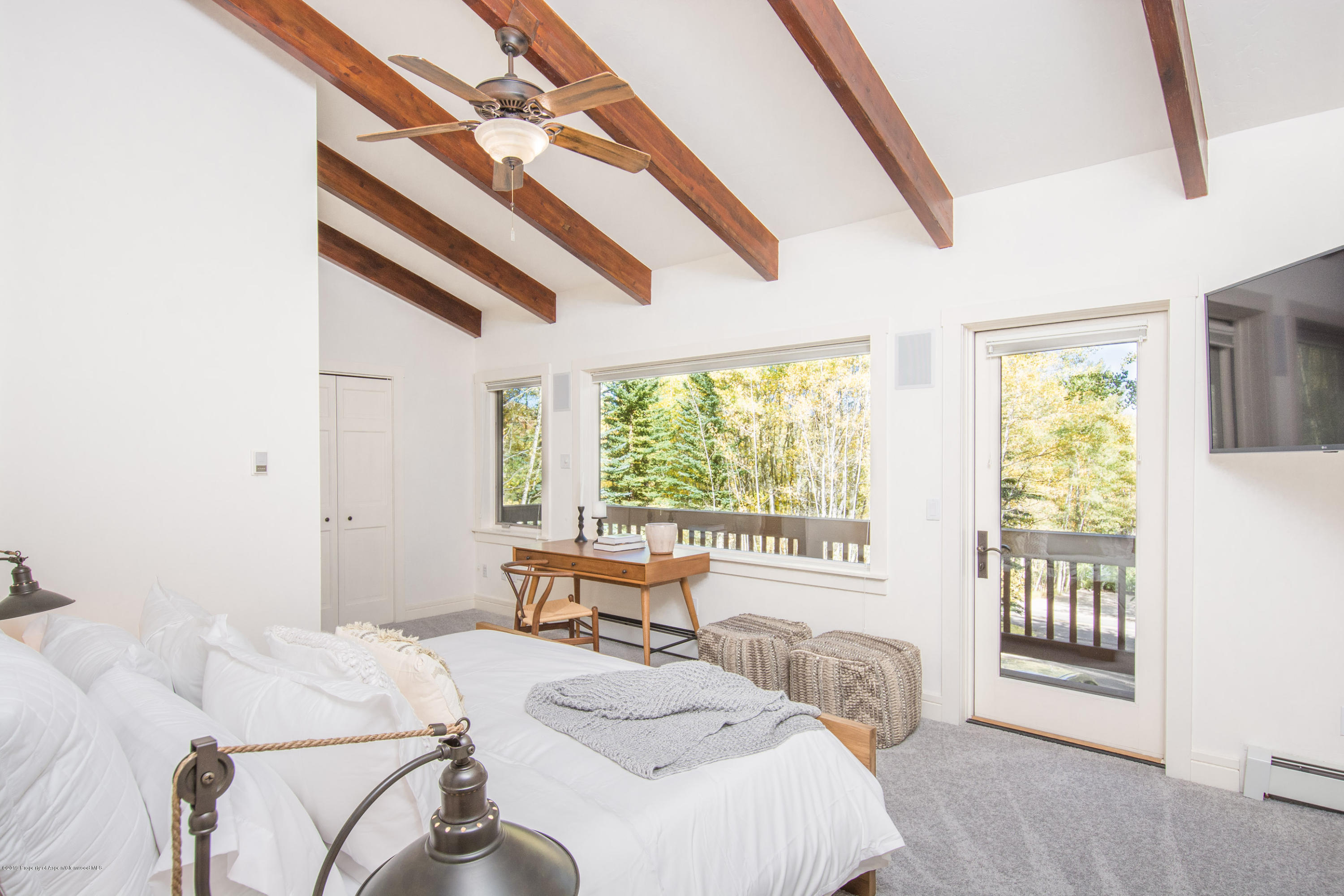 Bedroom with exposed wood beams, white bed, and windows overlooking trees.