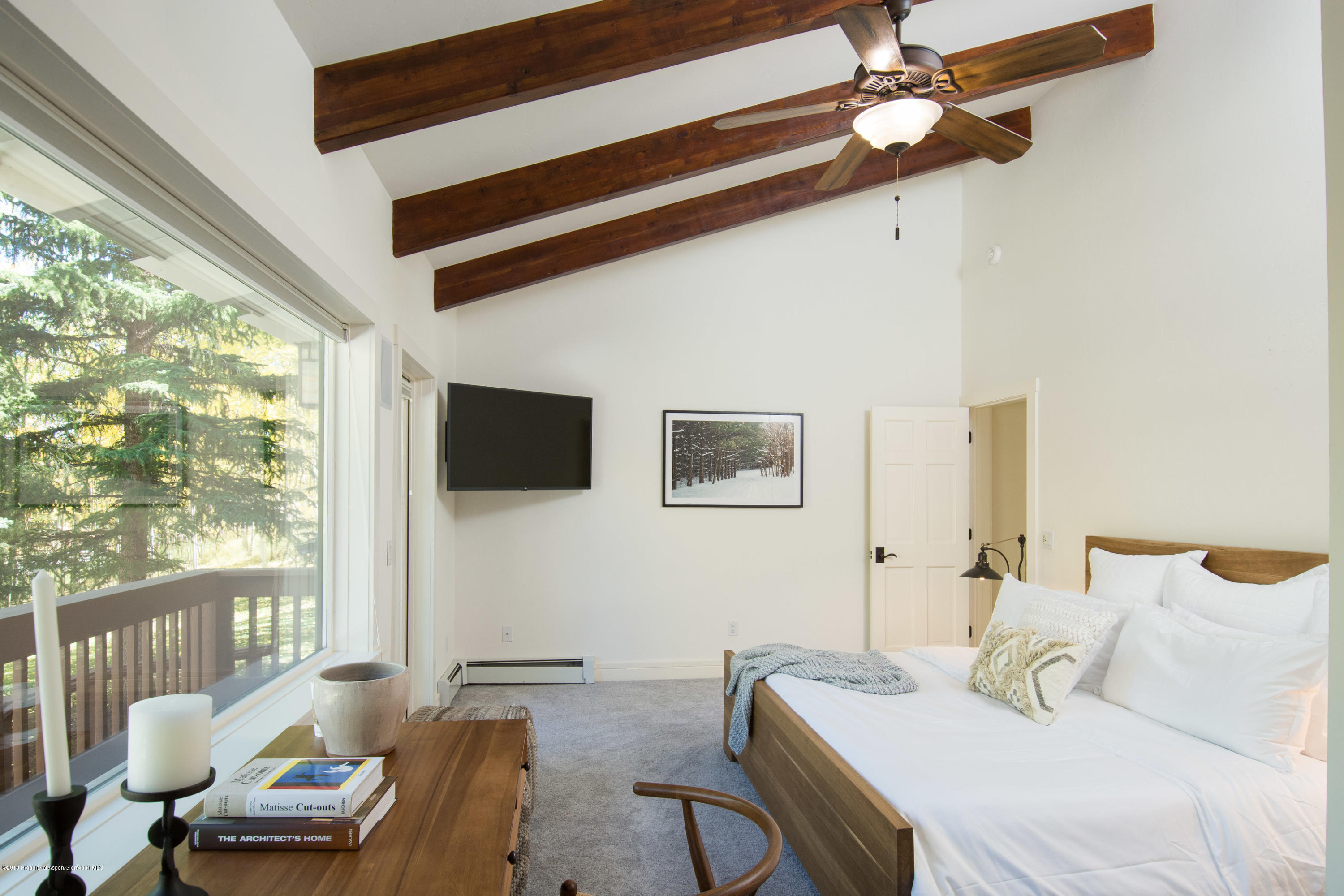 Bedroom with exposed wood ceiling beams, a large window overlooking evergreens, and a wood bed with white bedding.