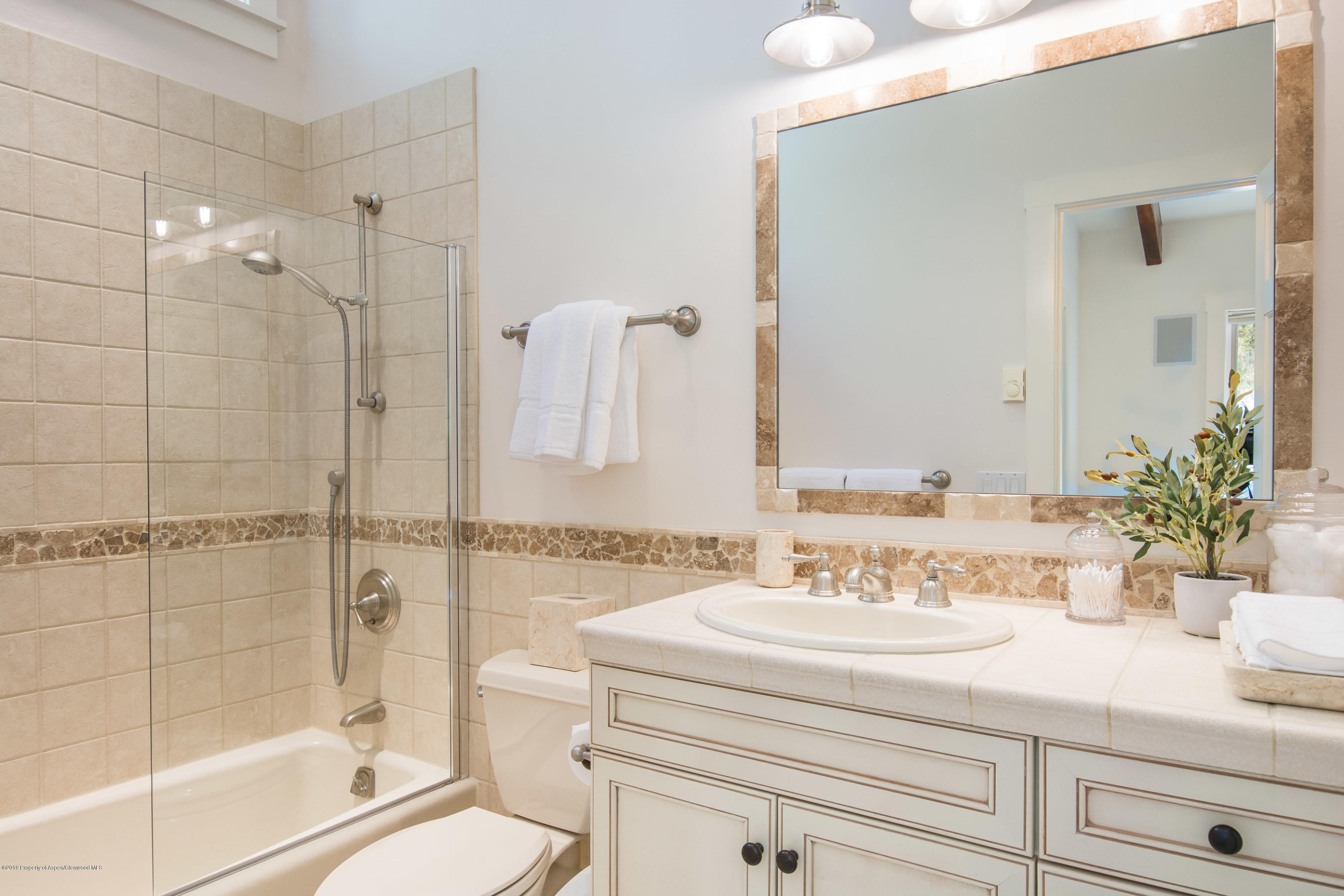 Bathroom with beige stone tile, glass shower enclosure, and white vanity beneath a large mirror.